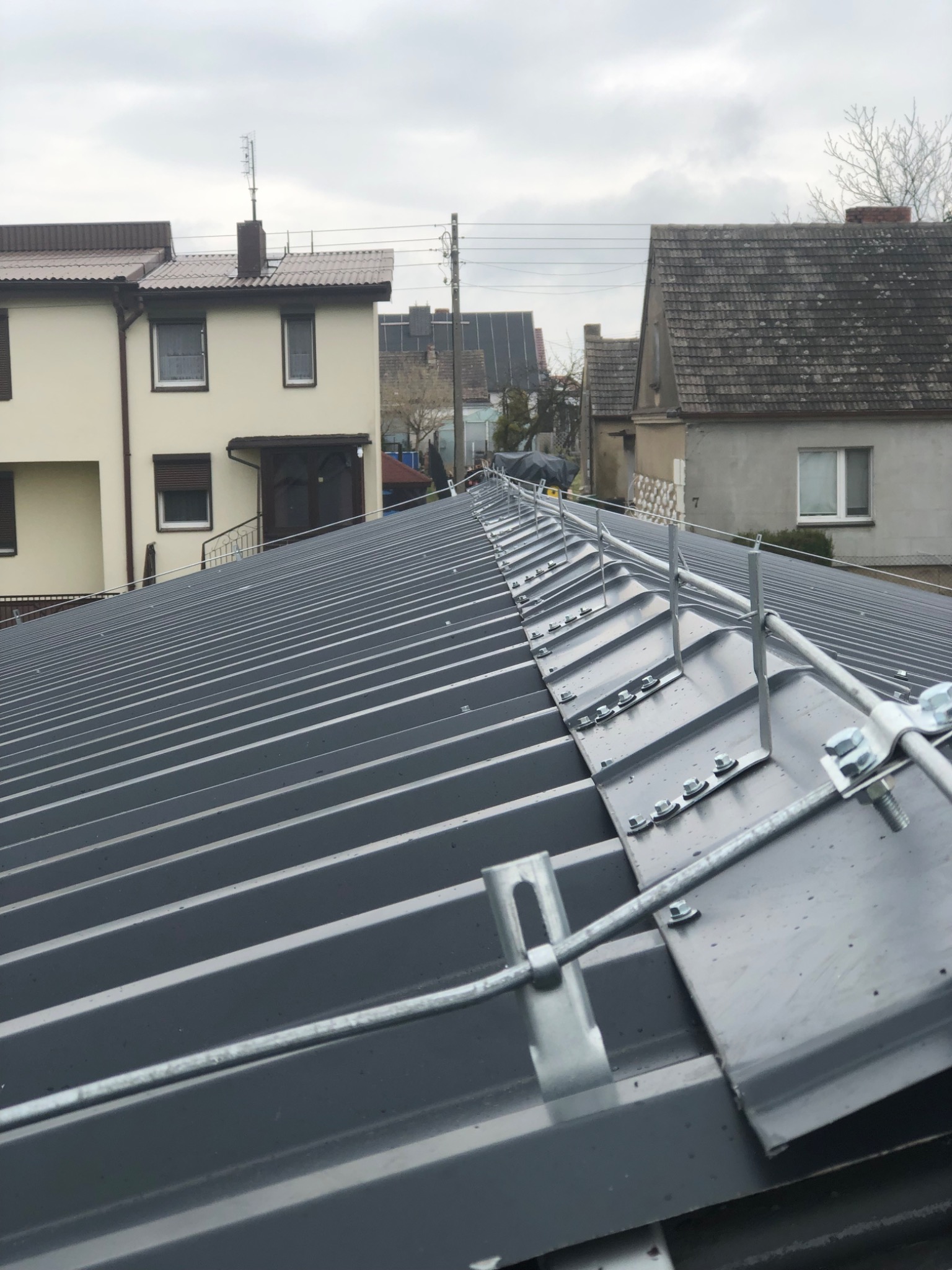 Szary dach z blachy trapezowej z zamontowaną instalacją odgromową, widoczne uchwyty i przewody, w tle zabudowa jednorodzinna w pochmurny dzień.