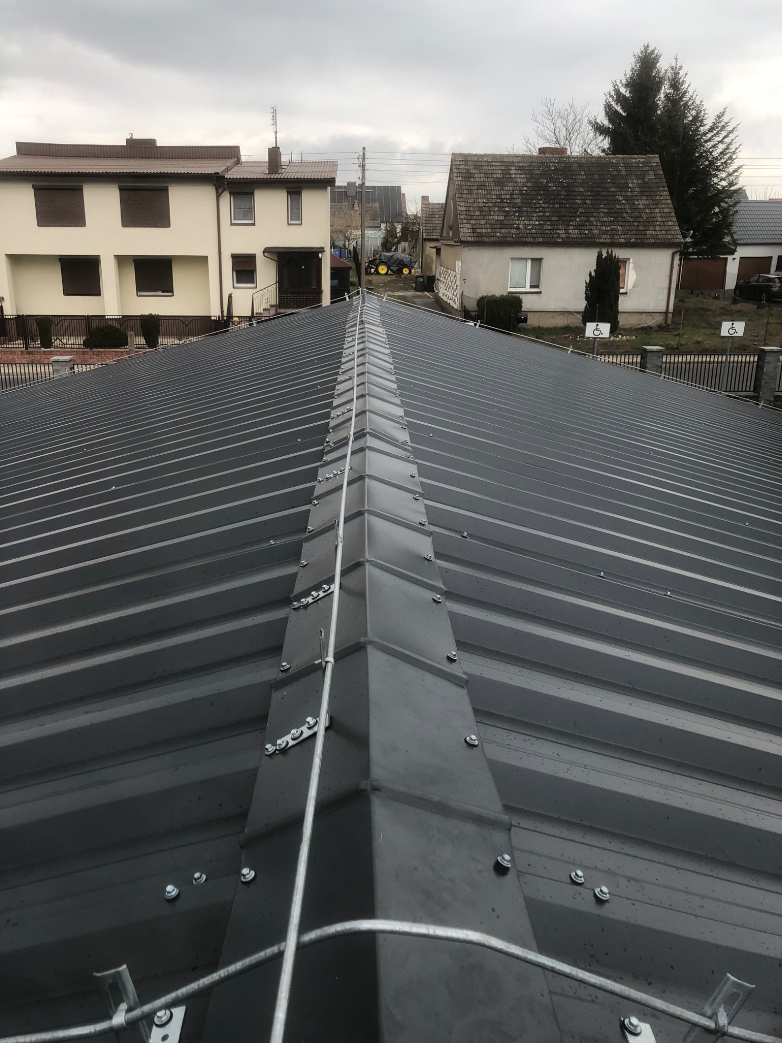 Widok z góry na nowo zainstalowaną, ciemnoszarą, metalową blachodachówkę z widocznym systemem odgromowym, na tle zabudowań jednorodzinnych w pochmurny dzień.