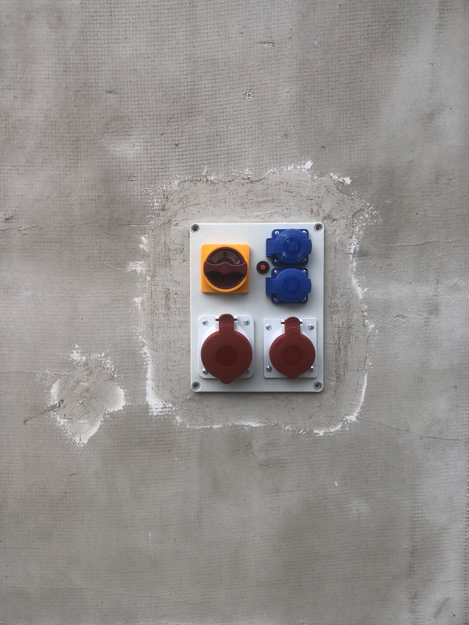 Panel elektryczny z wyłącznikiem i gniazdami przemysłowymi w kolorach czerwonym i niebieskim, zamontowany na surowej, szarej ścianie z widocznymi śladami po tynku.