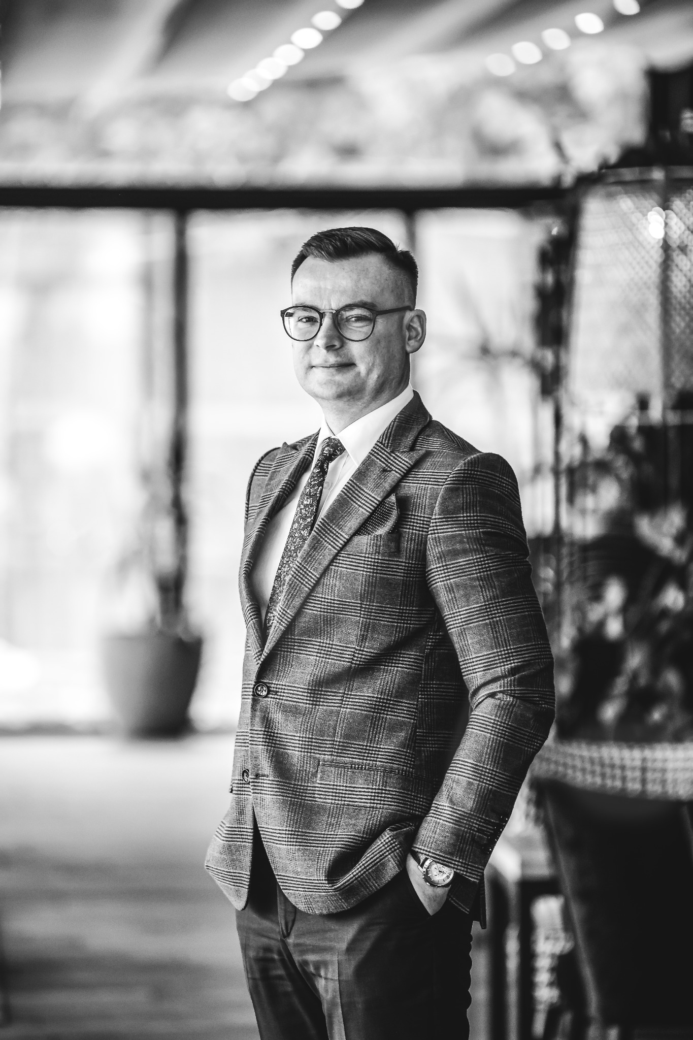 Portret mężczyzny w okularach i garniturze w kratę, z zegarkiem na ręce, stojącego na tle rozmytego wnętrza z roślinami, w czerni i bieli.