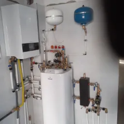 Kompletna instalacja grzewcza z piecem dwufunkcyjnym, zbiornikiem i naczyniami wyrównawczymi w pomieszczeniu technicznym, widoczne liczne rury i zawory.