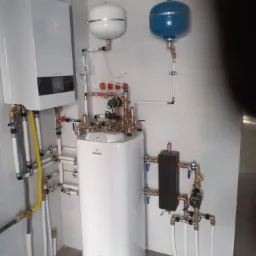 Biały zasobnik ciepłej wody użytkowej Galmet z licznymi rurami miedzianymi i białymi, zbiornik wyrównawczy w kolorze białym i niebieskim, zamontowane na ścianie z białych płytek, widoczne elementy...