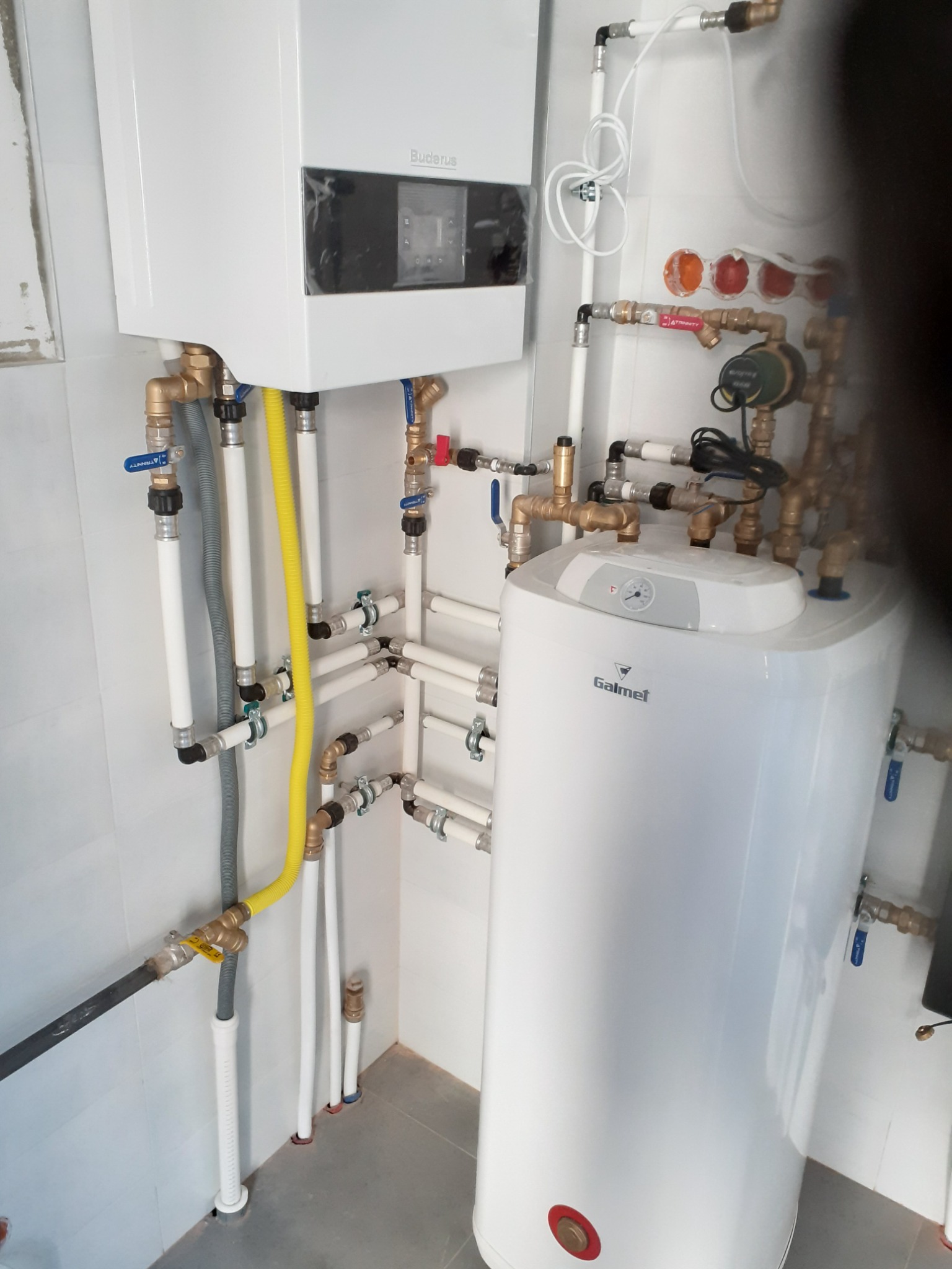 Biały piec Buderus i bojler Galmet połączone instalacją hydrauliczną z rurami w różnych kolorach w pomieszczeniu z szarą podłogą i białymi ścianami.