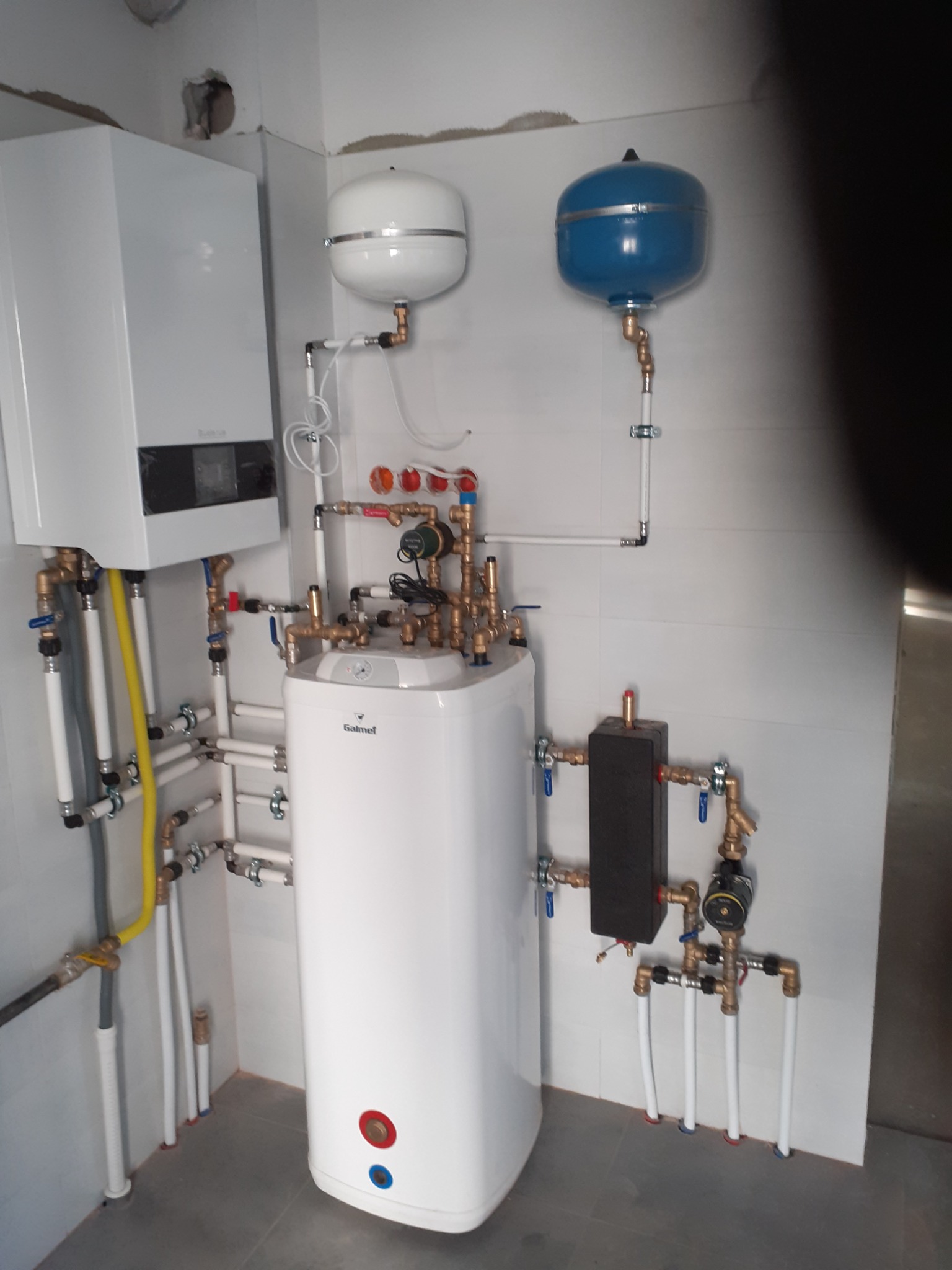 Biały zasobnik ciepłej wody użytkowej Galmet z licznymi rurami miedzianymi i białymi, zbiornik wyrównawczy w kolorze białym i niebieskim, zamontowane na ścianie z białych płytek, widoczne elementy...