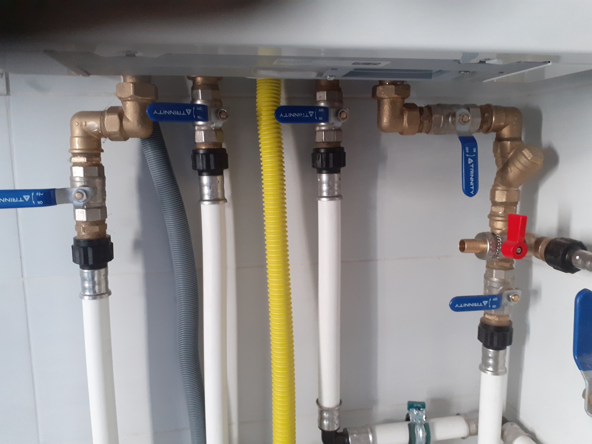 Złożona instalacja hydrauliczna z rurami z tworzywa, mosiężnymi złączkami i zaworami kulowymi z niebieskimi rączkami w białej szafce, widoczna żółta rura karbowana.