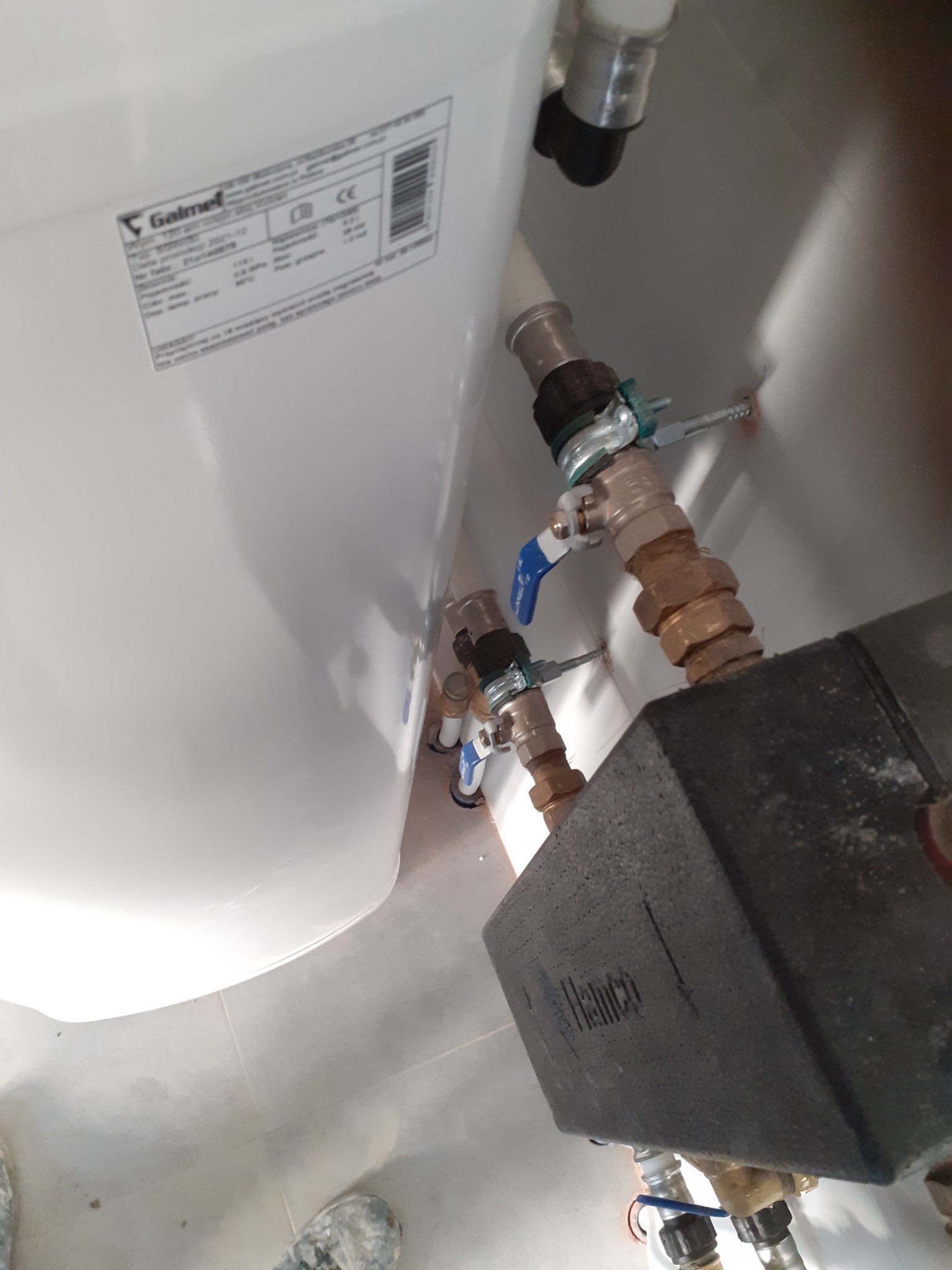 Biały bojler Galmet z podłączonymi miedzianymi rurami i zaworami z niebieskimi uchwytami, widoczny czarny element izolacyjny Henco, ujęcie od dołu z fragmentem butów na podłodze.