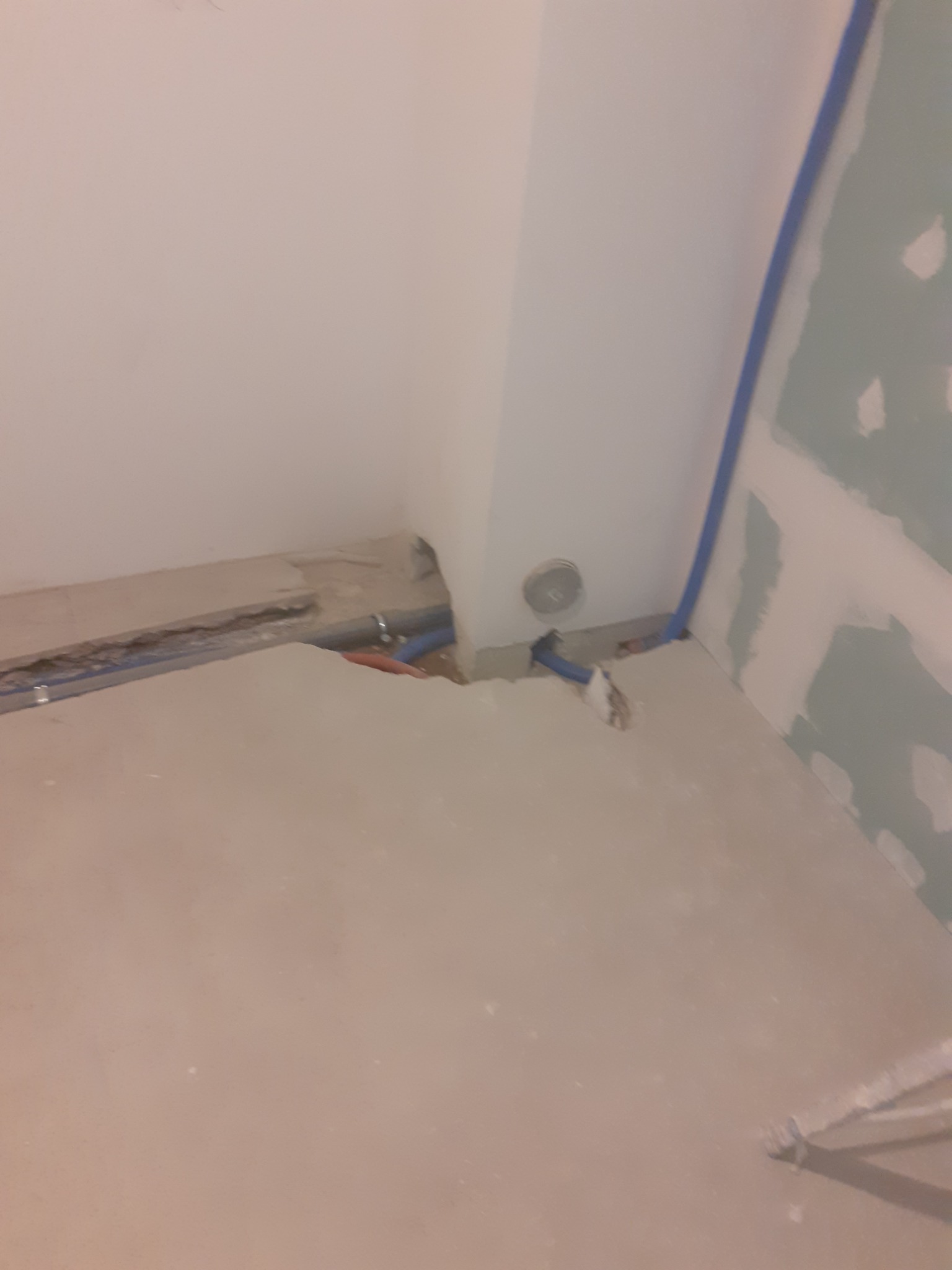 Niedokończona instalacja hydrauliczna z niebieskimi rurami w bruzdach w ścianie i podłodze, widoczna puszka instalacyjna w rogu pomieszczenia, ściana z płyt gipsowo-kartonowych w trakcie...