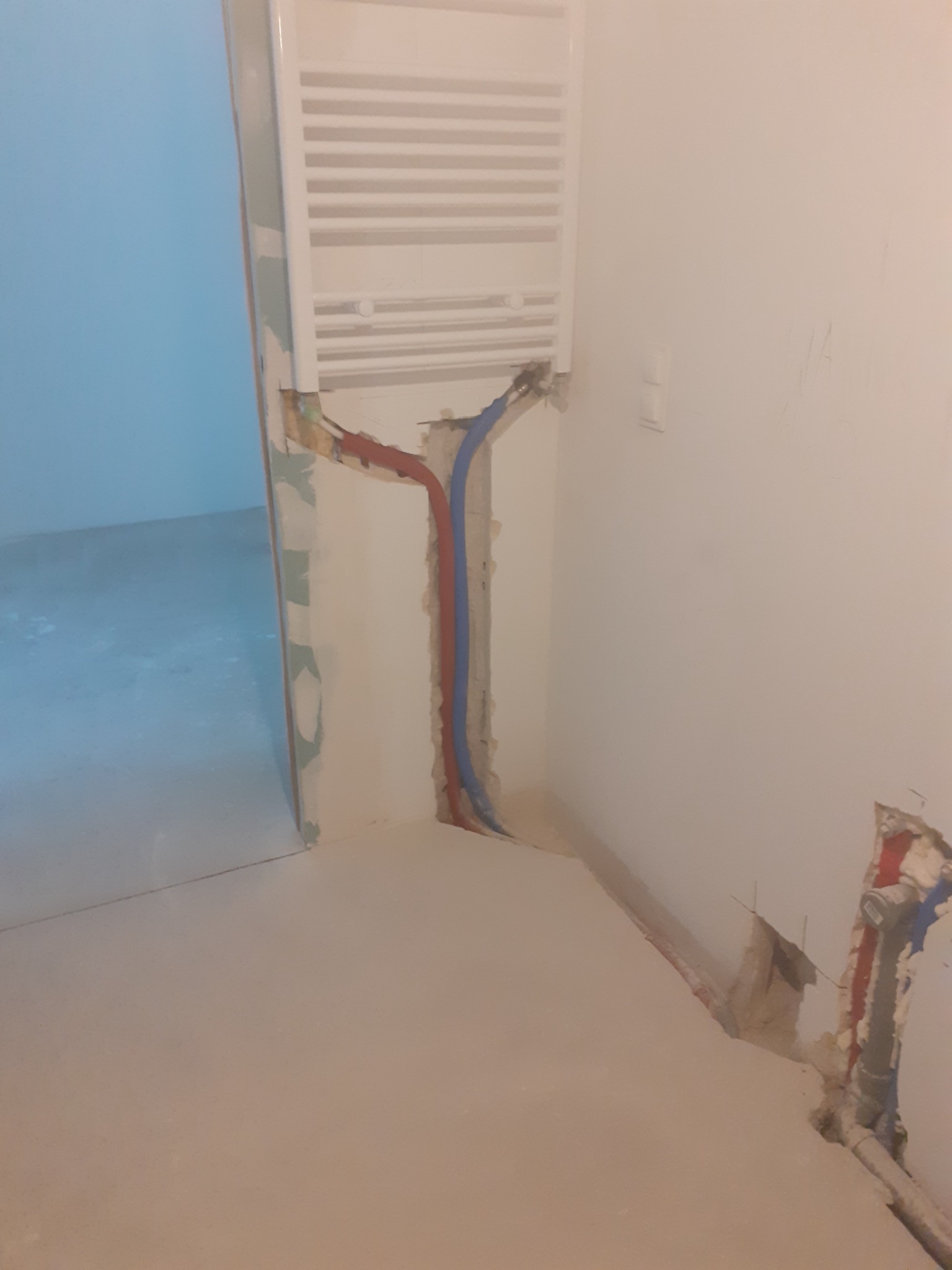 Biały grzejnik łazienkowy zamontowany na ścianie z widocznymi rurami miedzianymi i plastikowymi wystającymi ze ściany, w trakcie prac wykończeniowych.