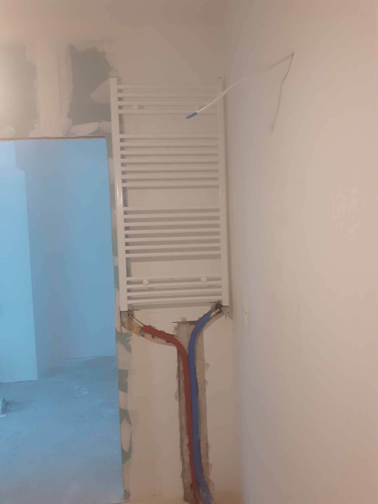 Biały grzejnik drabinkowy zamontowany na ścianie w trakcie remontu, z widocznymi rurami instalacji hydraulicznej w kolorach czerwonym i niebieskim, wystającymi ze ściany, na tle surowych ścian...
