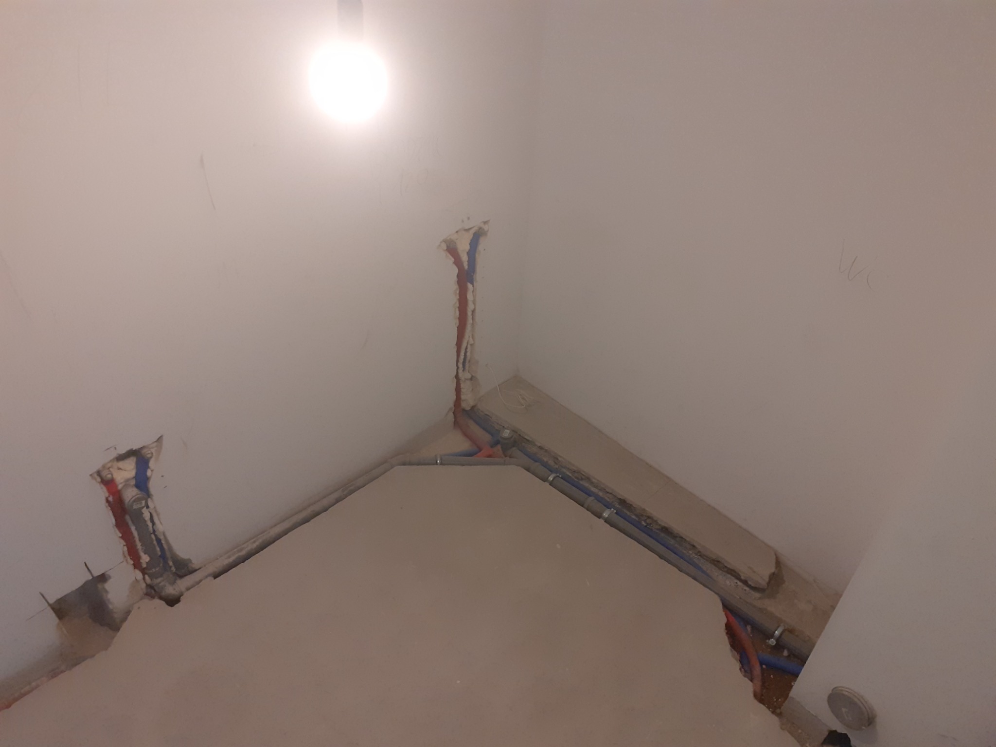 Fragment ściany w trakcie remontu z widocznymi przewodami instalacji hydraulicznej, w tym rury czerwone i niebieskie, oraz ślady po kuciu w ścianie i podłodze.