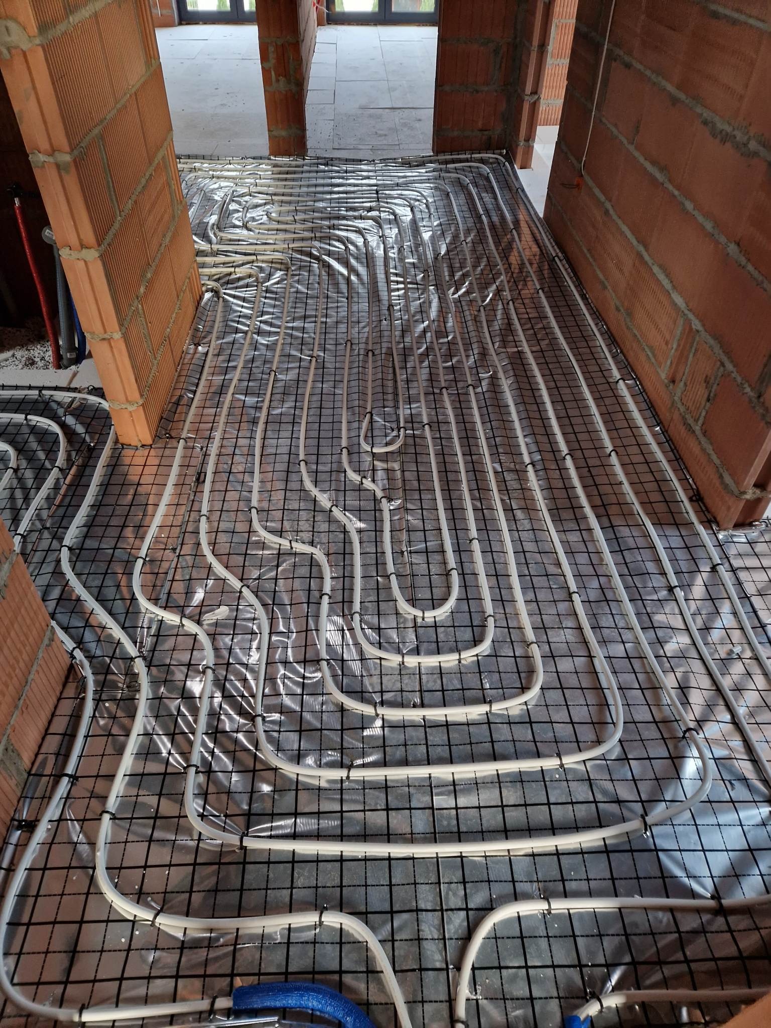 Białe rury ogrzewania podłogowego ułożone na czarnej siatce, widoczne ceglane ściany w surowym stanie.