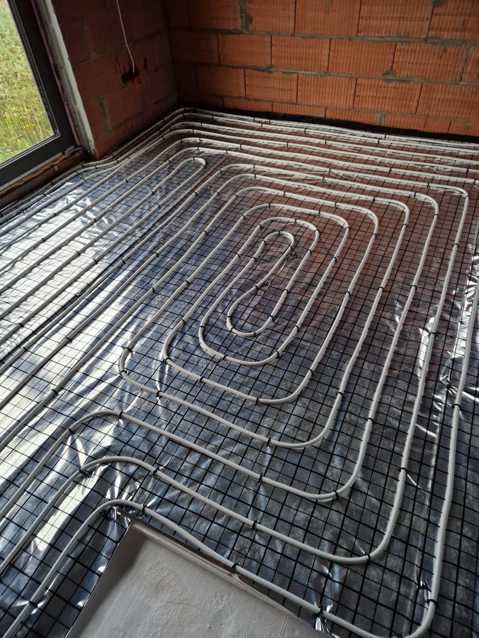 Białe rurki systemu ogrzewania podłogowego rozłożone na folii z czarną siatką, widok z góry na surowe ściany z cegły.