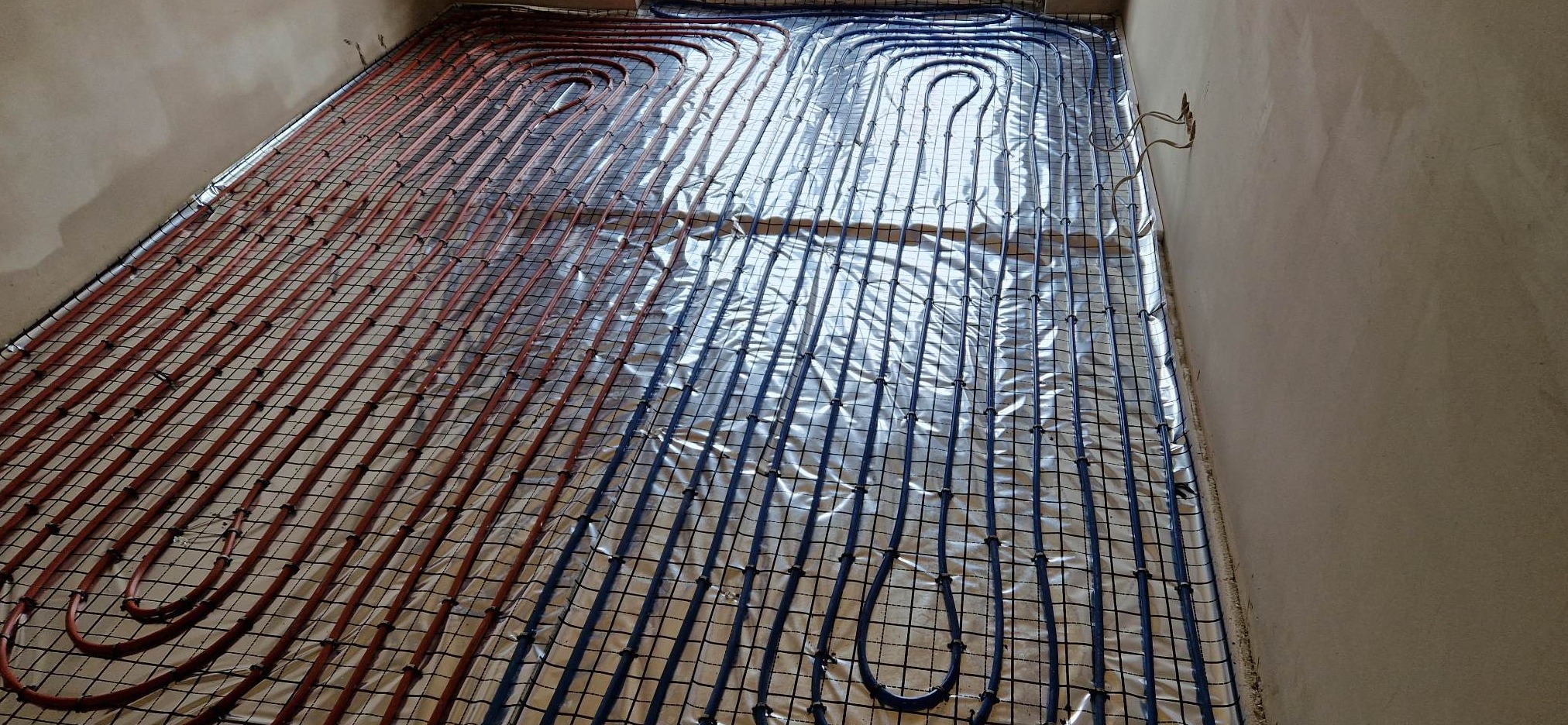Instalacja ogrzewania podłogowego z rurami w kolorach czerwonym i niebieskim, ułożonymi na folii izolacyjnej i przymocowanymi do siatki montażowej.