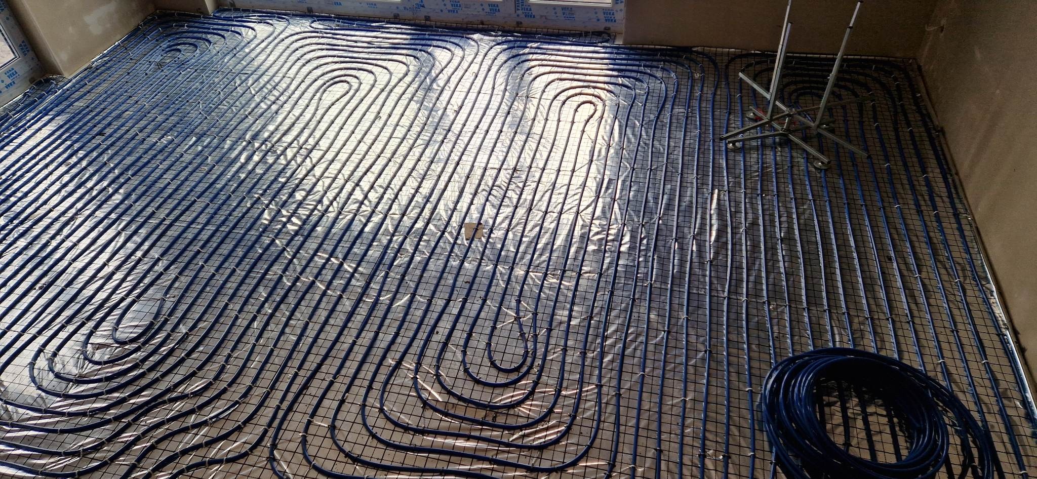 Niebieskie rury systemu ogrzewania podłogowego ułożone na folii odblaskowej i siatce, widoczny fragment zwiniętej rury oraz stojak montażowy w rogu pomieszczenia.