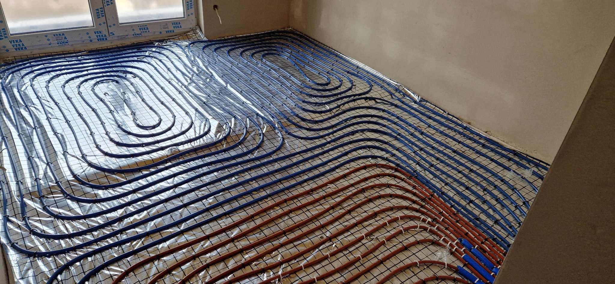 Instalacja rur grzewczych, częściowo w kolorze niebieskim i pomarańczowym, ułożonych na srebrnej folii odblaskowej i czarnej siatce w nowym budynku, widoczne okna z logo VEKA.