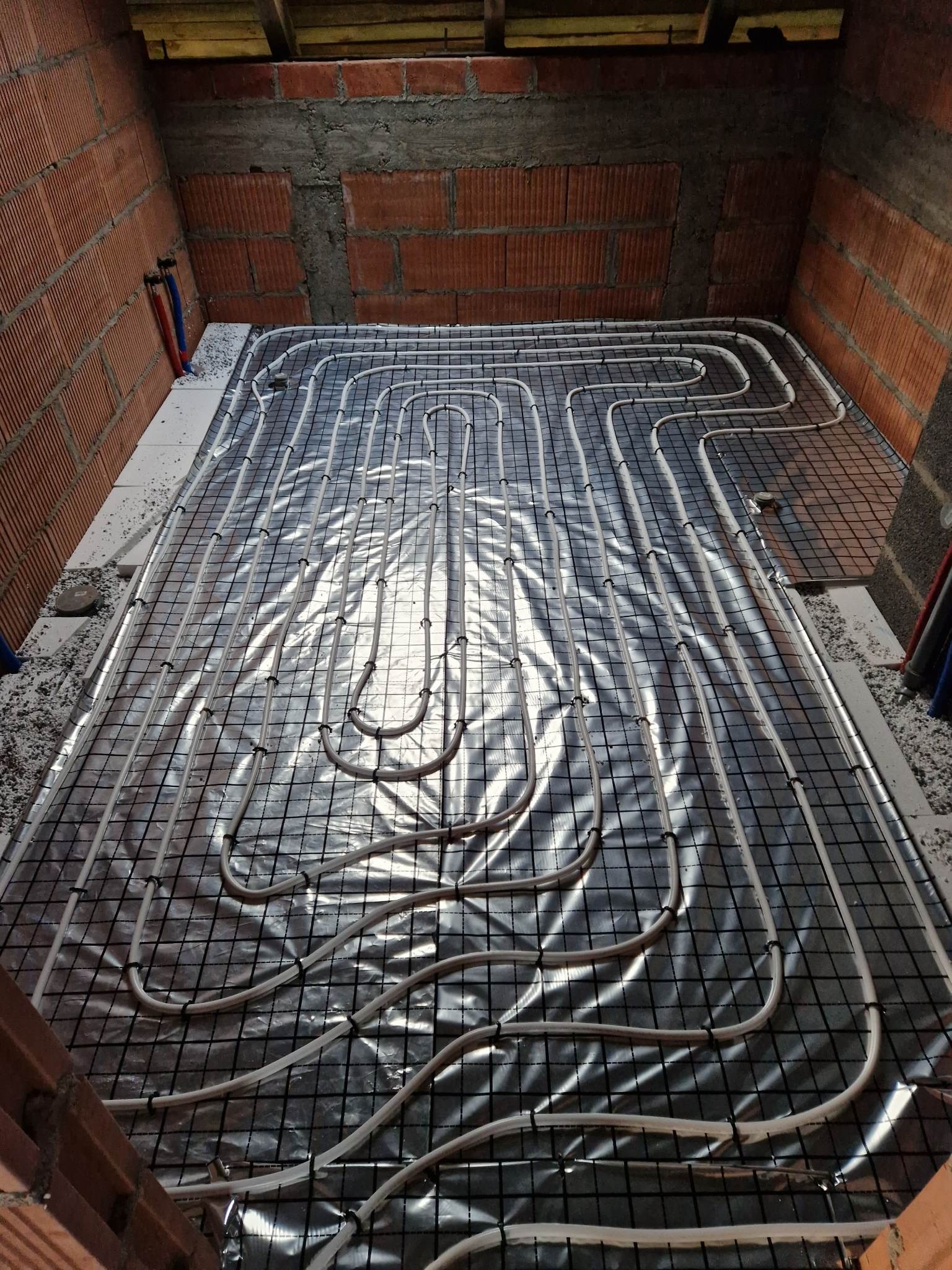 Instalacja ogrzewania podłogowego w surowym stanie, rury grzewcze zamocowane do siatki na folii izolacyjnej, ściany z czerwonej cegły.