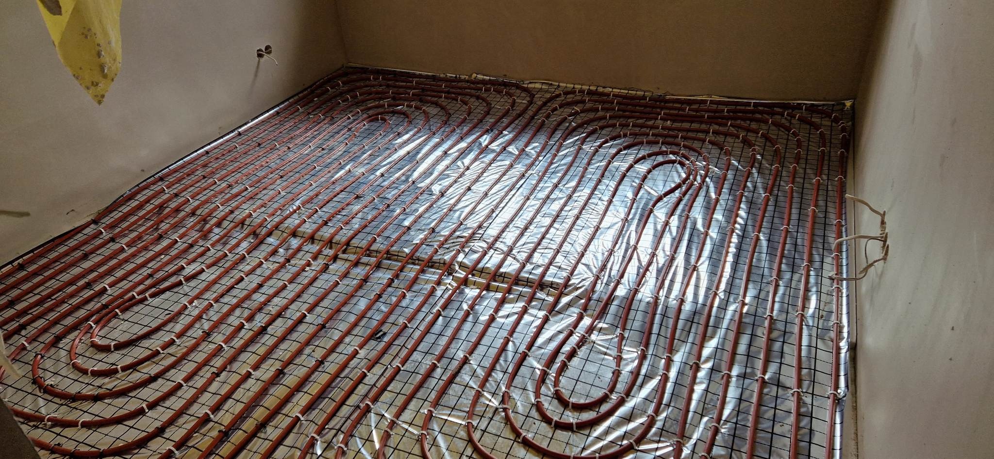 Instalacja ogrzewania podłogowego z rurami w kolorze ceglastym, rozłożonymi na folii izolacyjnej i siatce montażowej, widoczne mocowania rur do siatki.