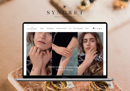 Laptop wyświetla stronę internetową sklepu jubilerskiego Syncret z trzema modelkami prezentującymi pierścionki z diamentami, leżący na kamiennej płycie otoczonej złotą biżuterią.