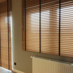 dominik florczak - Drewniane żaluzje poziome w jasnym odcieniu drewna z czarnymi taśmami na oknach w pokoju z grzejnikiem pod oknem.
