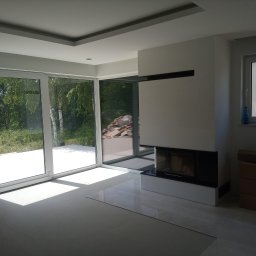 dominik florczak - Jasne, przestronne wnętrze w trakcie wykańczania, z dużymi przesuwnymi oknami wychodzącymi na zieleń, nowoczesnym kominkiem z czarnymi akcentami i częściowo ułożonymi płytkami podłogowymi.