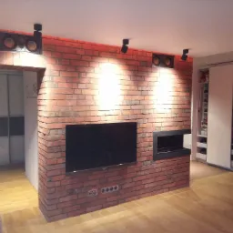 Salon z ceglaną ścianą, na której zawieszony jest telewizor i wbudowany kominek elektryczny, oświetlenie punktowe na suficie, widoczna zabudowa szafy przesuwnej w tle.