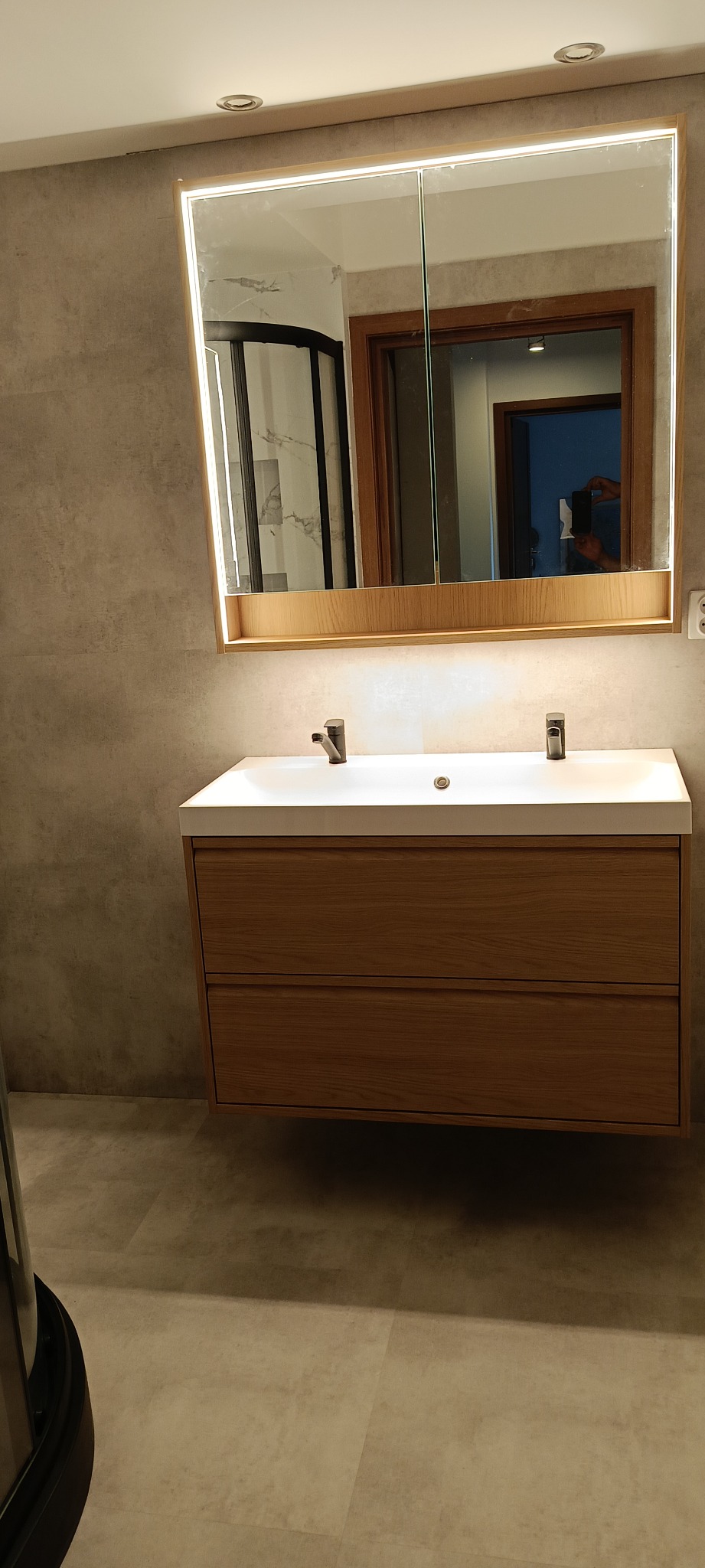 Nowoczesna łazienka z podświetlanym lustrem szafkowym, białą umywalką nablatową z drewnianą szafką podwieszaną i szarą płytką imitującą beton na ścianach.