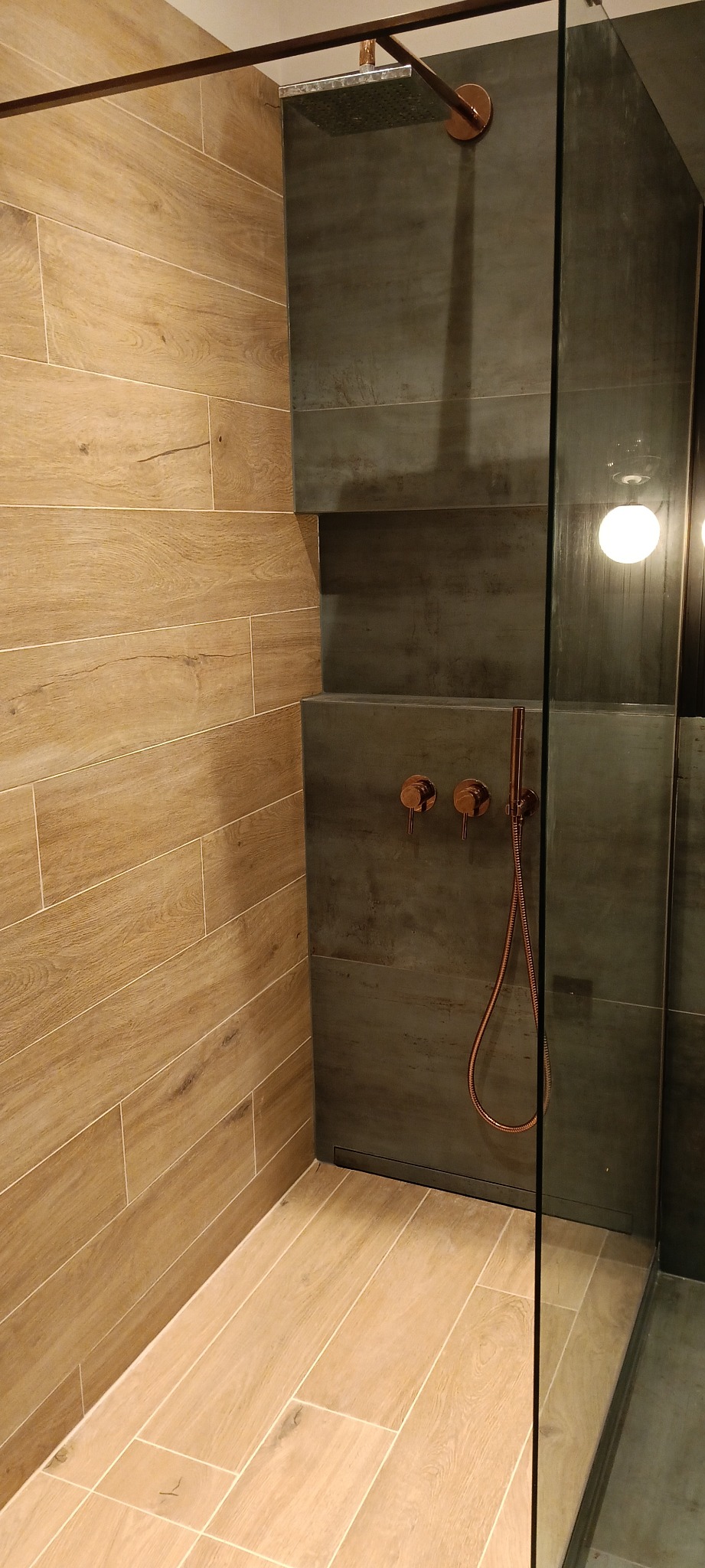 Nowoczesna kabina prysznicowa z okładziną imitującą drewno i szarymi płytkami, armatura w kolorze miedzi, widoczny odpływ liniowy.