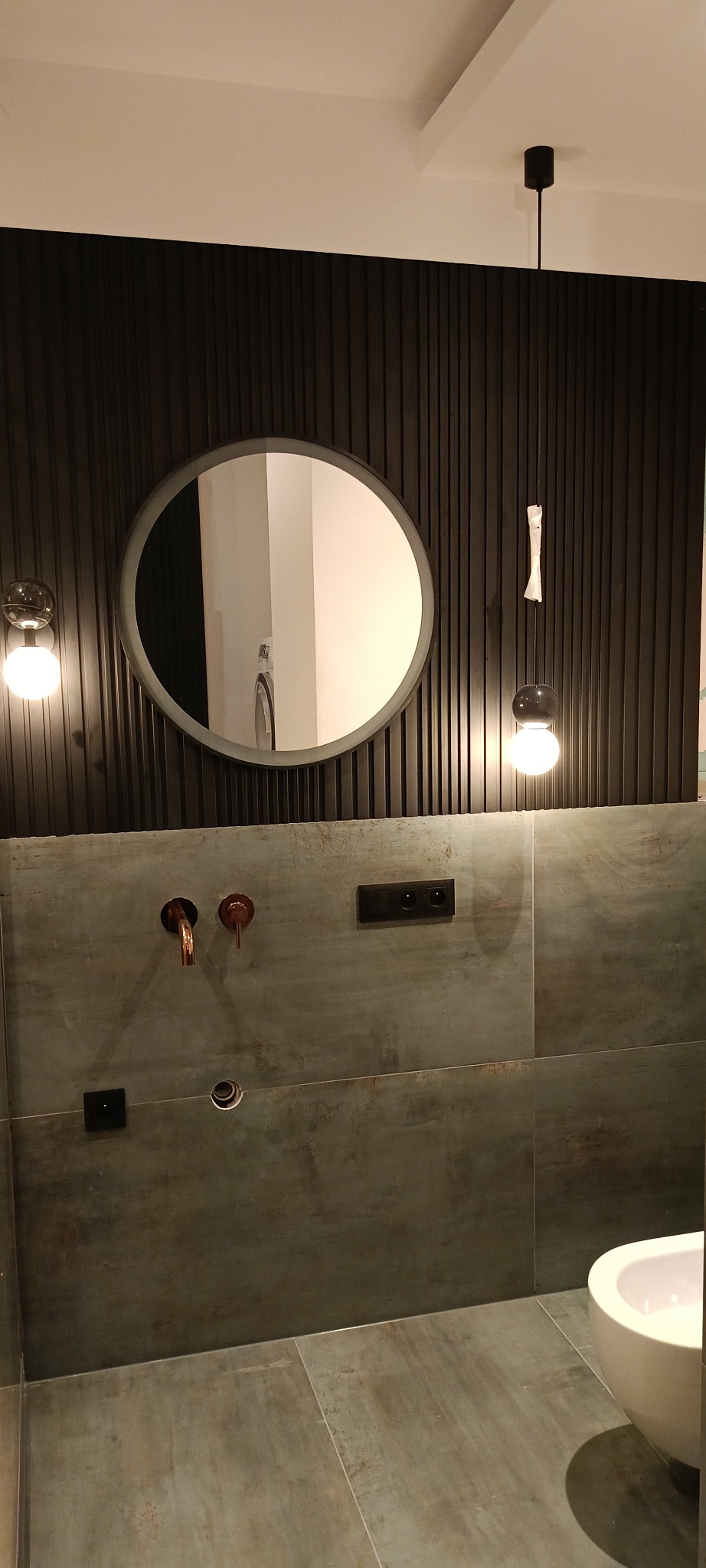 Nowoczesna łazienka z okrągłym lustrem podświetlanym, czarną ścianą z pionowych paneli, miedzianą armaturą i szarą płytką imitującą beton.