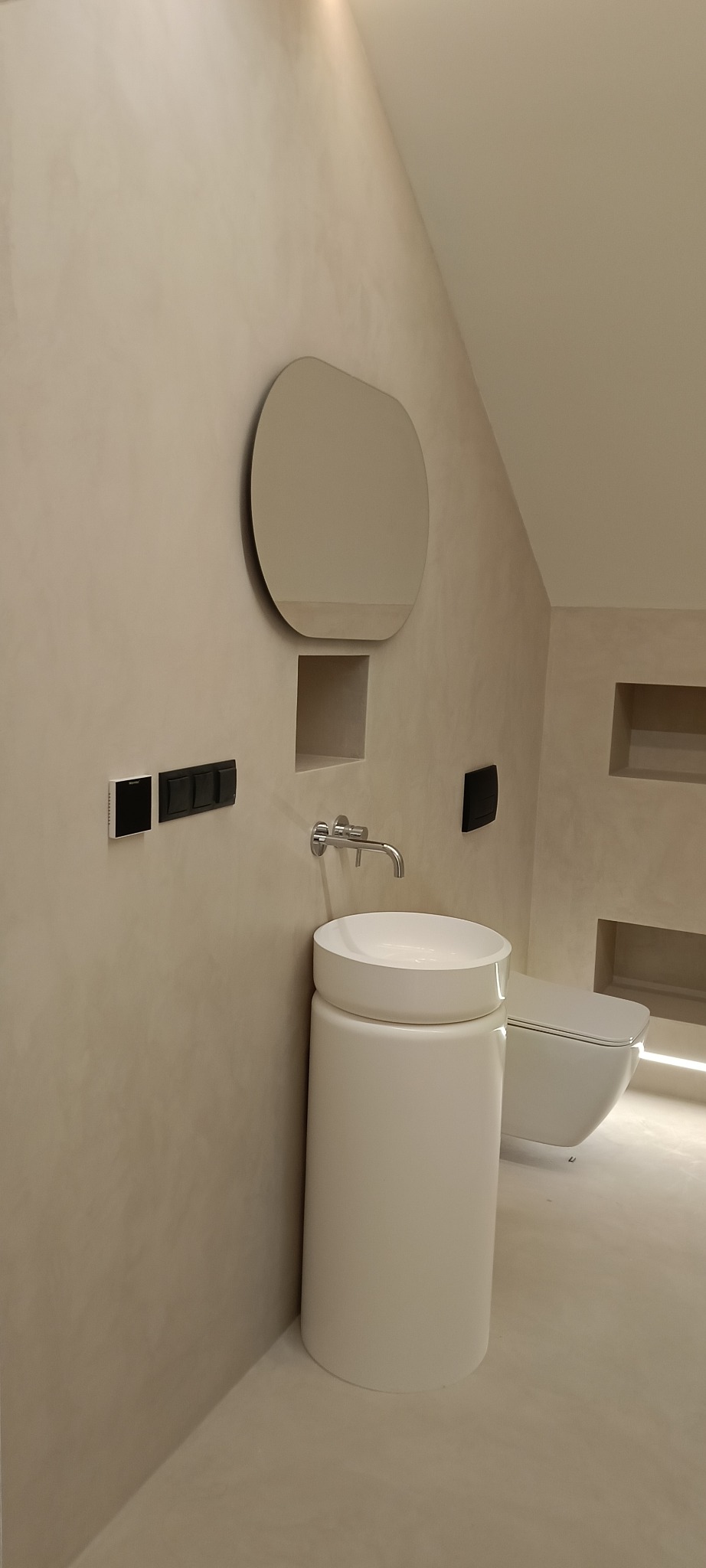 Minimalistyczna łazienka w Łodzi z beżowymi ścianami, okrągłym lustrem, umywalką na postumencie i podwieszaną toaletą z podświetleniem LED w niszy.