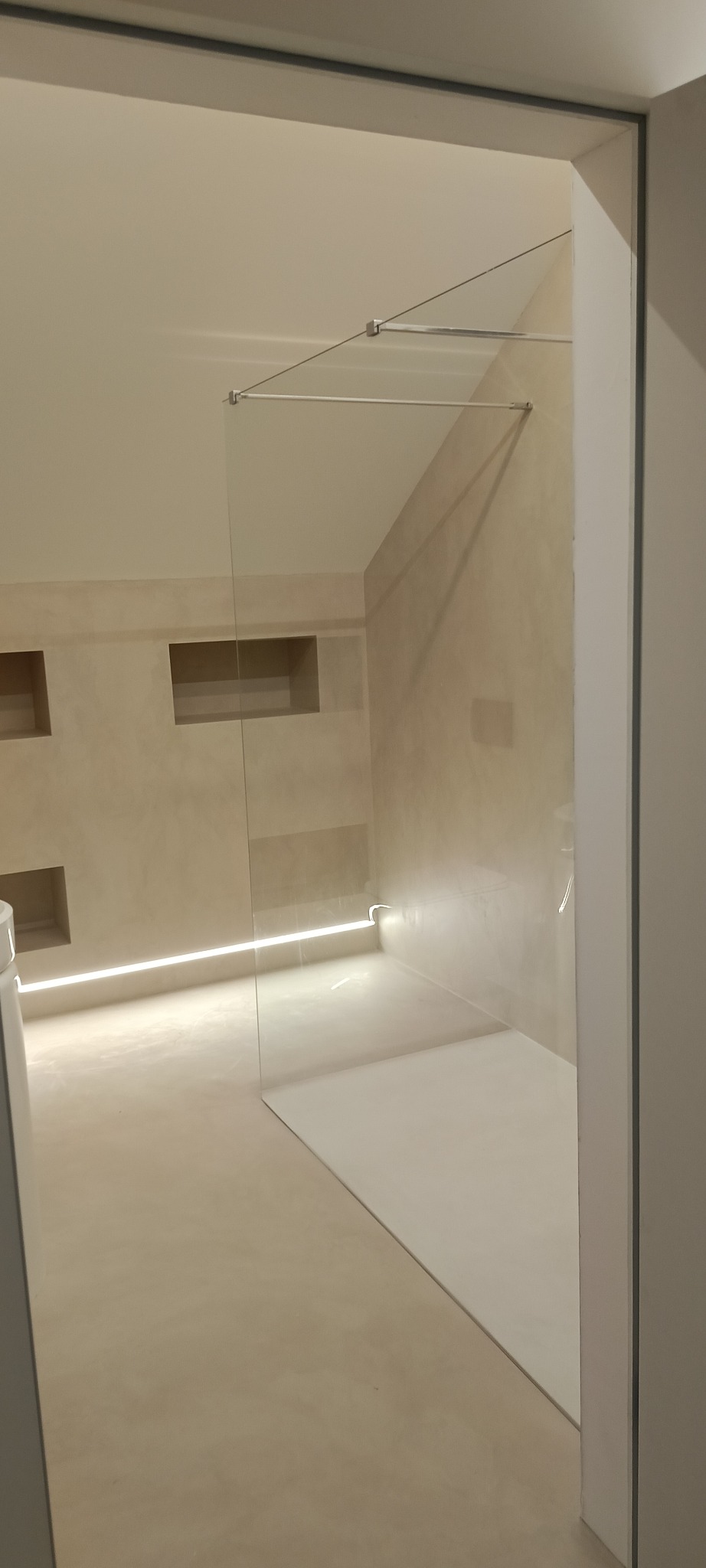 Nowoczesna kabina prysznicowa ze szklaną ścianką, oświetleniem LED wzdłuż podłogi i wnękami w ścianie wykończonej beżowym tynkiem.