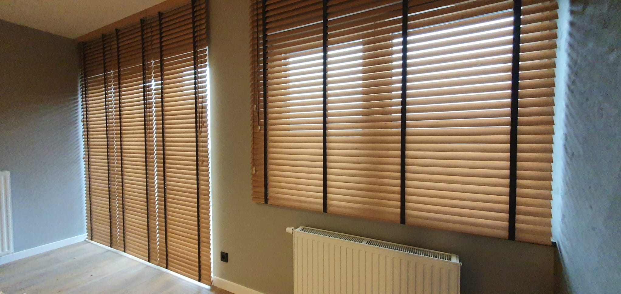 Drewniane żaluzje poziome w jasnym odcieniu drewna z czarnymi taśmami na oknach w pokoju z grzejnikiem pod oknem.