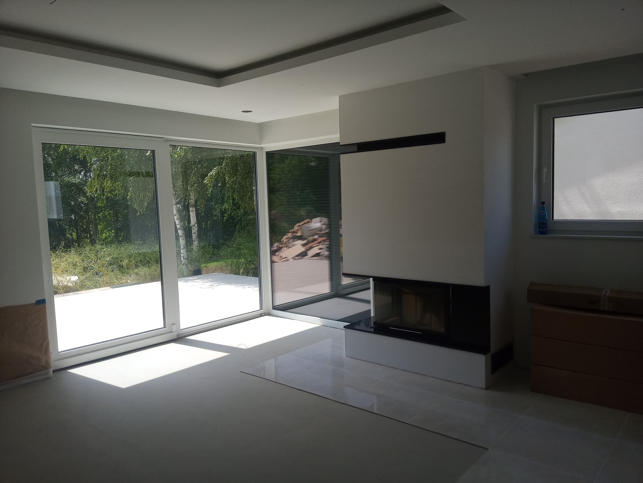 Jasne, przestronne wnętrze w trakcie wykańczania, z dużymi przesuwnymi oknami wychodzącymi na zieleń, nowoczesnym kominkiem z czarnymi akcentami i częściowo ułożonymi płytkami podłogowymi.