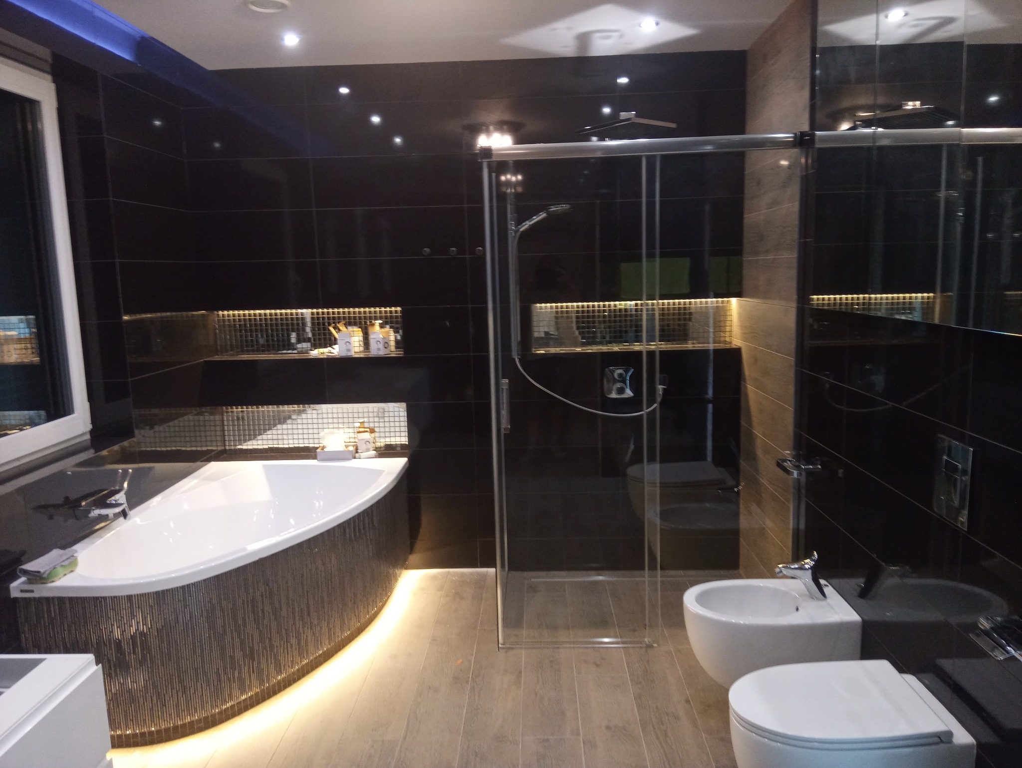 Nowoczesna łazienka z czarnymi płytkami, podświetlaną wanną, kabiną prysznicową ze szklanymi drzwiami i drewnopodobną podłogą. Na półkach dekoracyjne akcesoria łazienkowe.