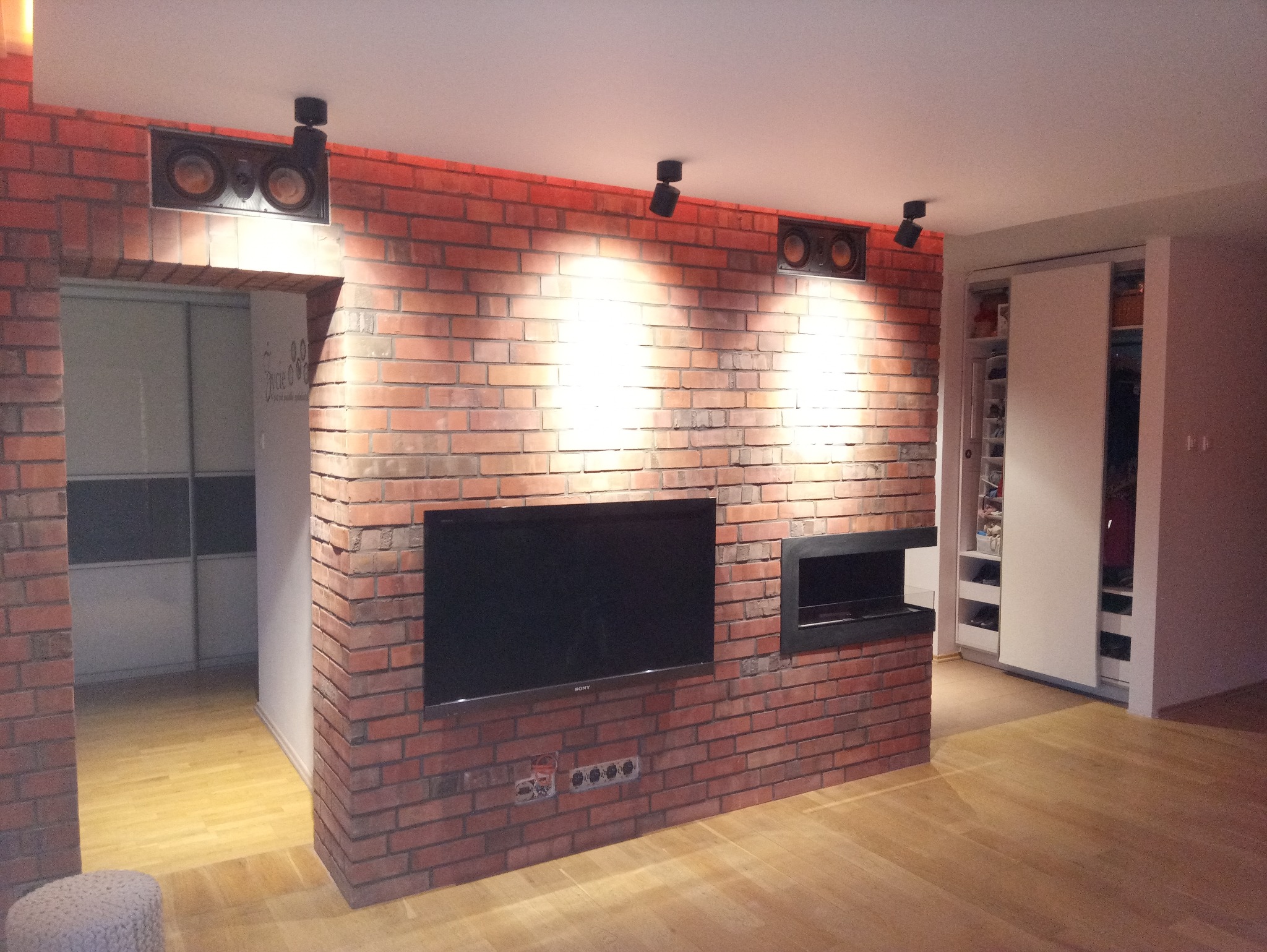 Salon z ceglaną ścianą, na której zawieszony jest telewizor i wbudowany kominek elektryczny, oświetlenie punktowe na suficie, widoczna zabudowa szafy przesuwnej w tle.