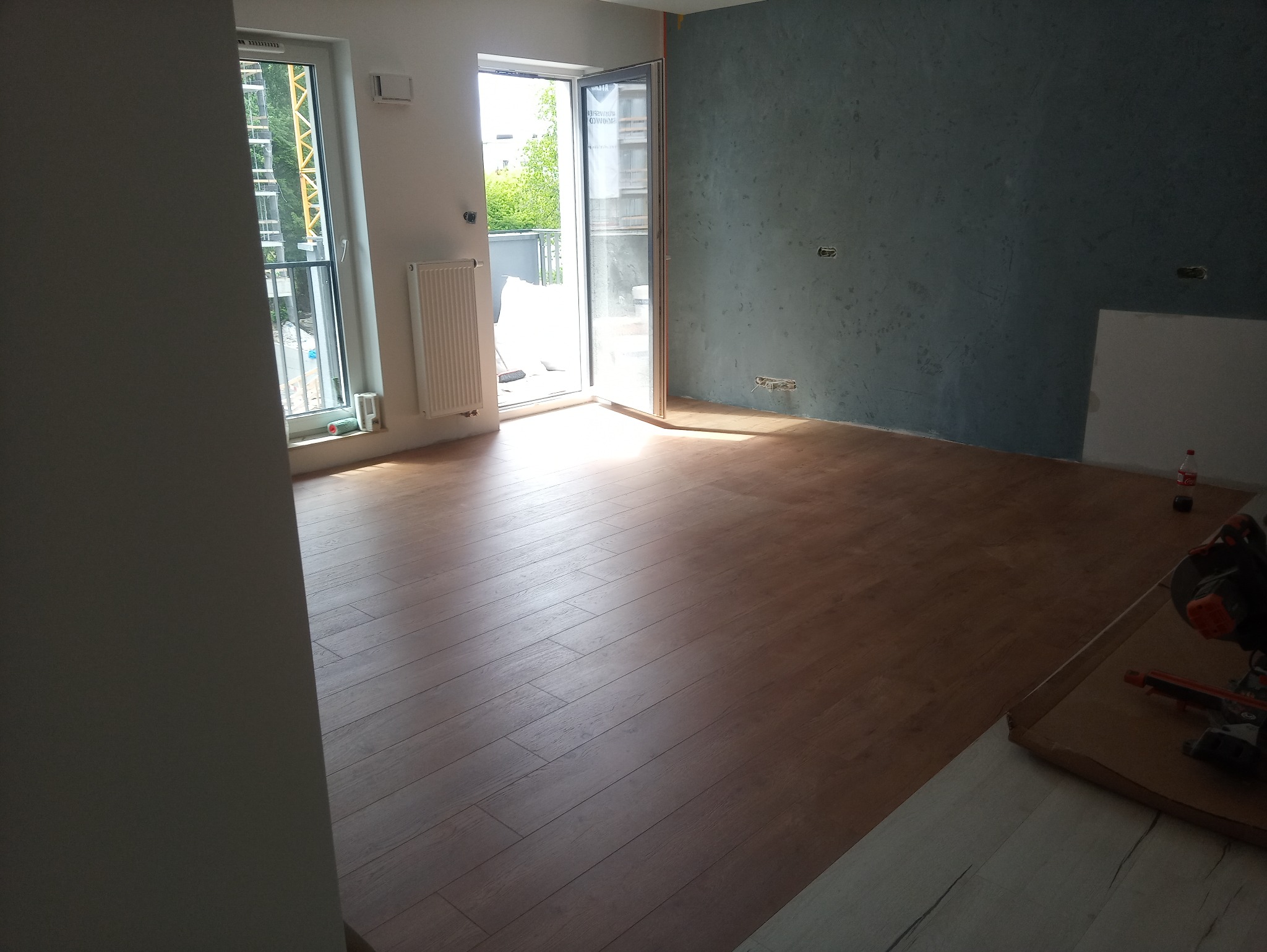 Nowe panele podłogowe w jasnym pokoju z otwartym balkonem, widoczna piła do paneli i ściana z dekoracyjną fakturą.