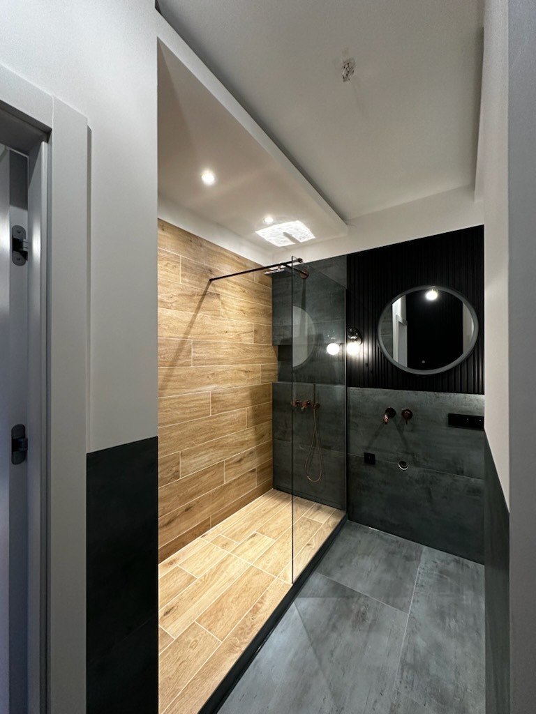 Nowoczesna łazienka z kabiną prysznicową typu walk-in, wykończona płytkami imitującymi drewno na ścianie i podłodze w strefie prysznica, oraz szarą płytką w pozostałej części, z czarnymi akcentami...