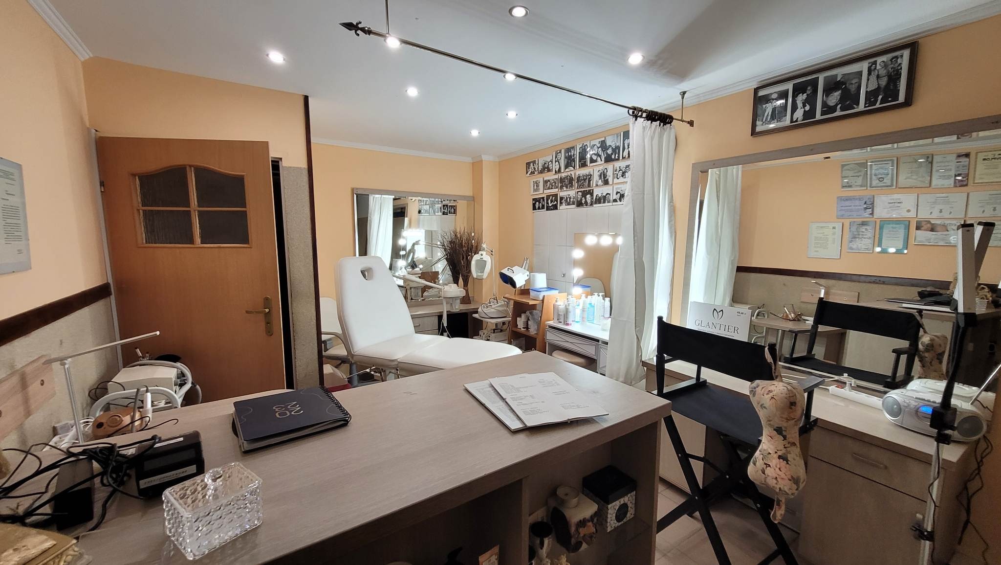 Jasne wnętrze gabinetu kosmetycznego z białym fotelem zabiegowym, biurkiem z dokumentami, lustrami i oświetleniem punktowym, dekorowane zdjęciami i certyfikatami na ścianach.