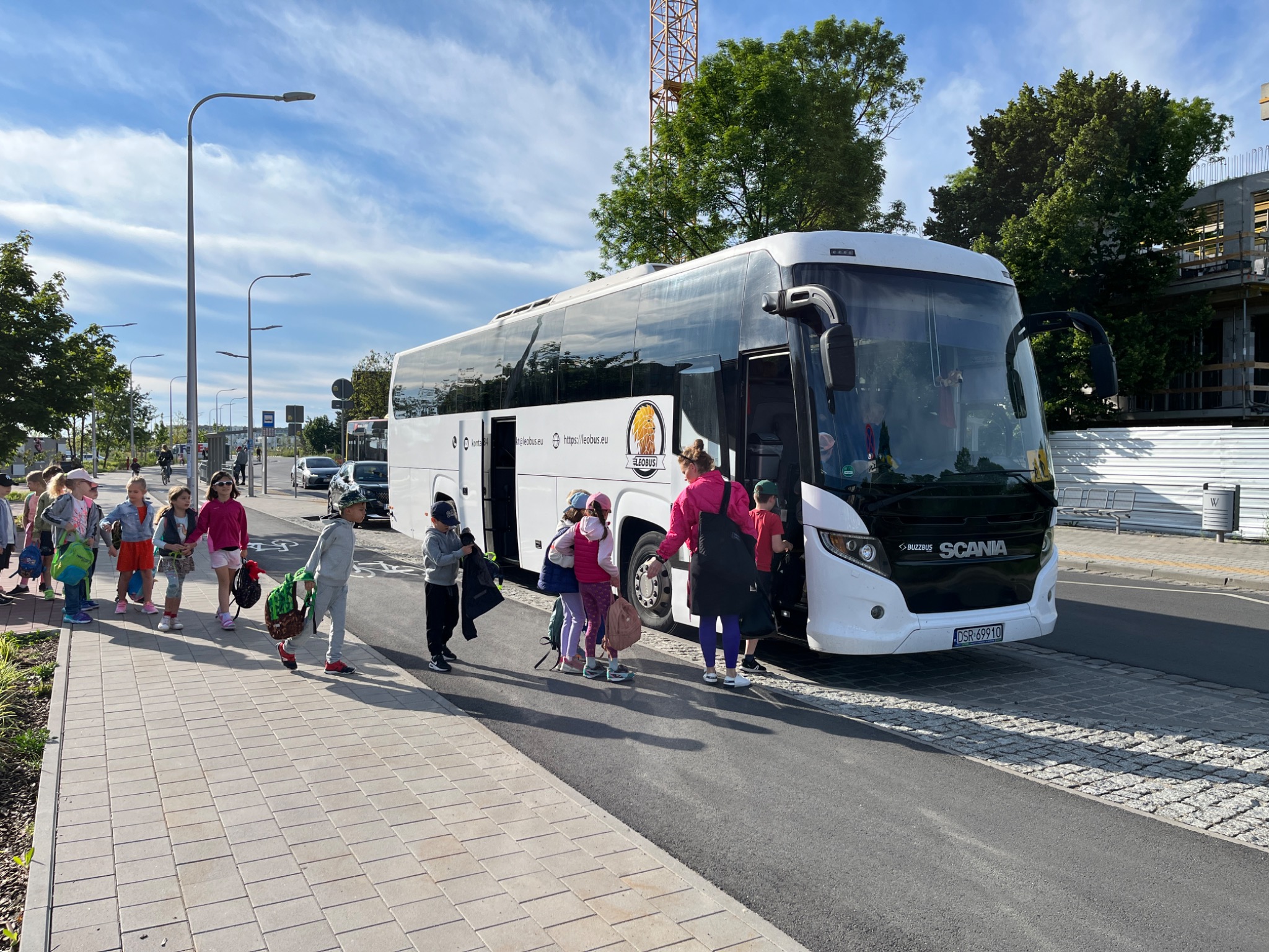 Grupa dzieci w różnym wieku wsiada do białego autokaru Scania z otwartymi drzwiami na tle miejskiego krajobrazu z budową w tle.