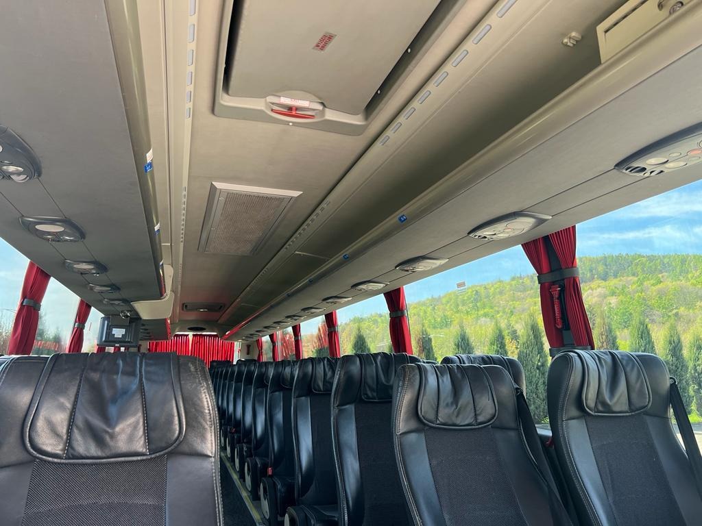 Wnętrze autobusu turystycznego z czarnymi skórzanymi siedzeniami, czerwonymi zasłonami i widokiem na zielone wzgórza za oknem.