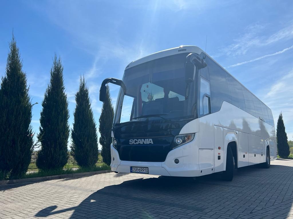 Biały autokar marki Scania z otwartymi drzwiami, zaparkowany na kostce brukowej, na tle niebieskiego nieba z delikatnymi chmurami i rzędu wysokich, zielonych drzew.