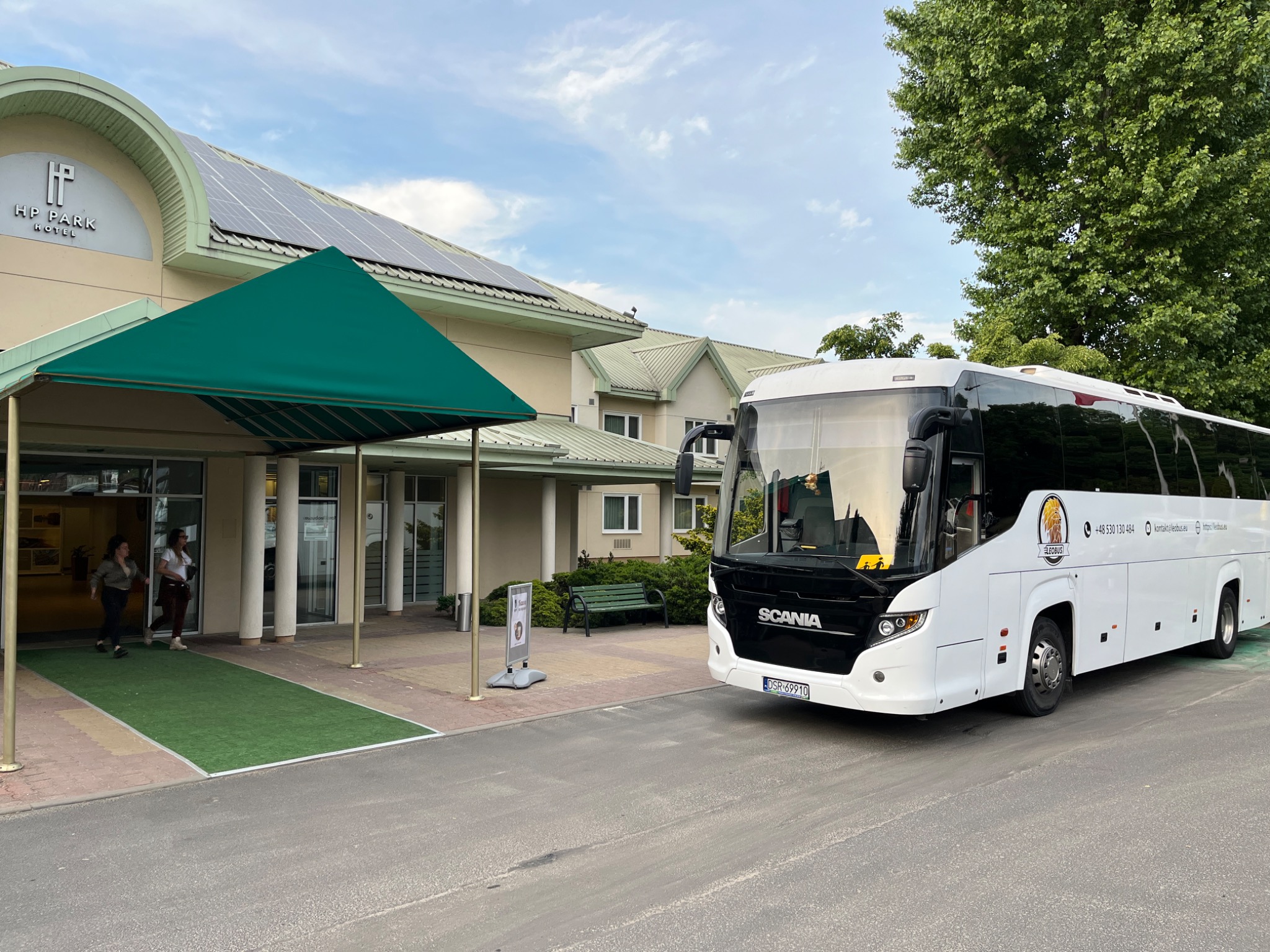 Biały autokar Scania zaparkowany przed hotelem HP Park z zielonym daszkiem nad wejściem i panelami słonecznymi na dachu. Na autokarze widoczne logo Leobus i dane kontaktowe.