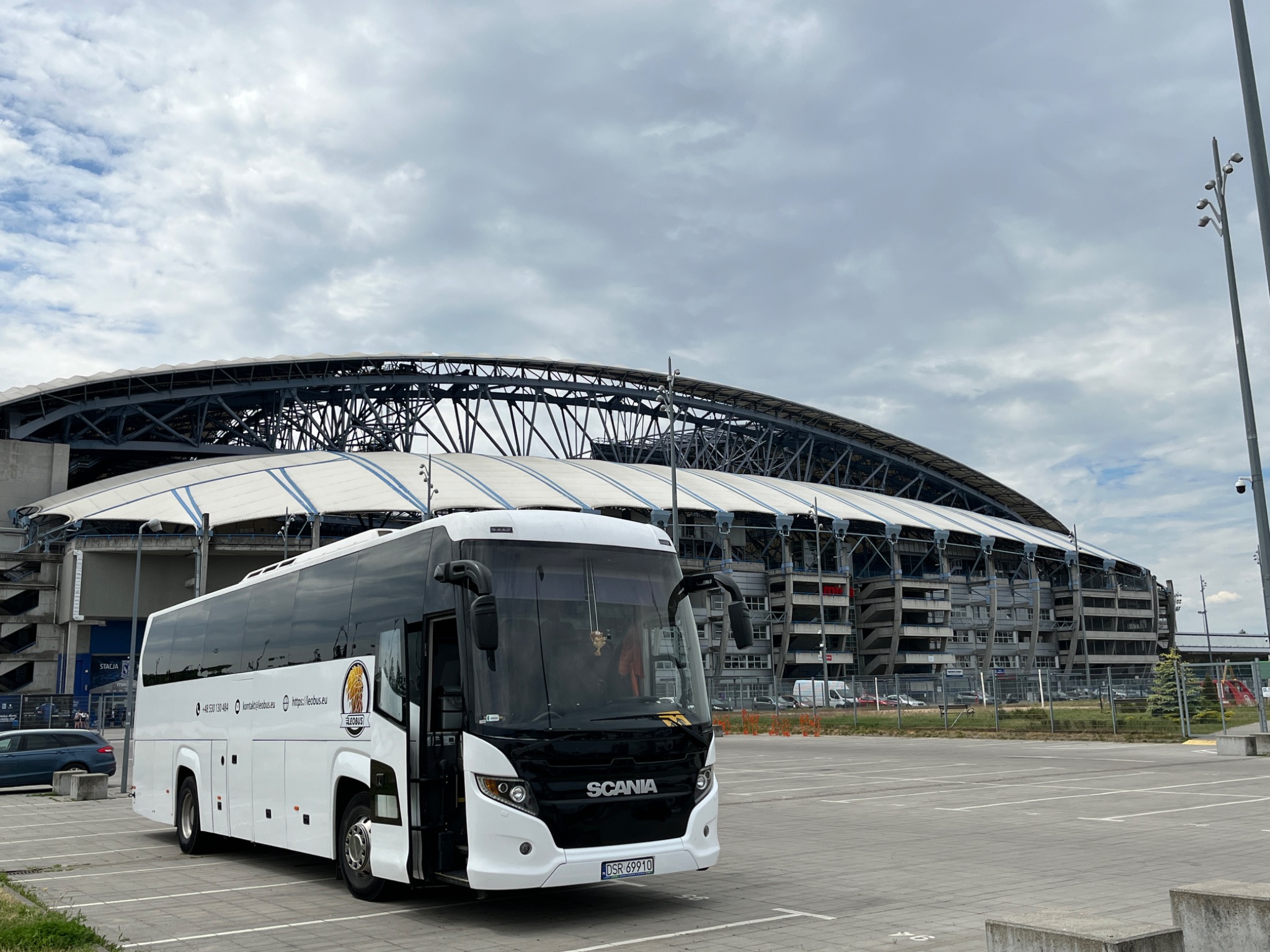 Biały autokar Scania z otwartymi drzwiami na parkingu przed stadionem miejskim, pochmurne niebo w tle.