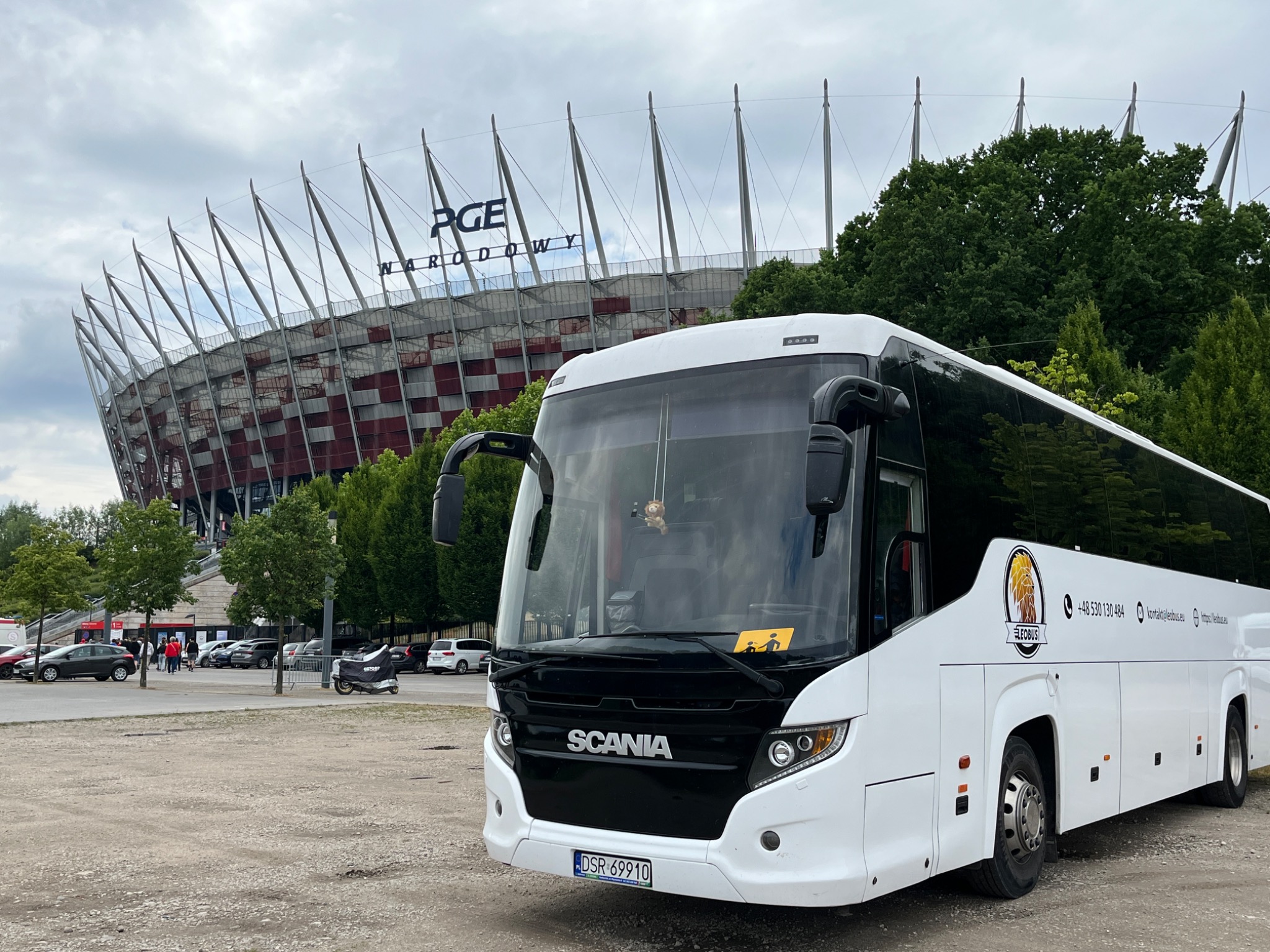 Biały autokar Scania z logiem firmy na tle Stadionu Narodowego w Warszawie, widoczny znak ostrzegawczy 'dzieci' na przedniej szybie.