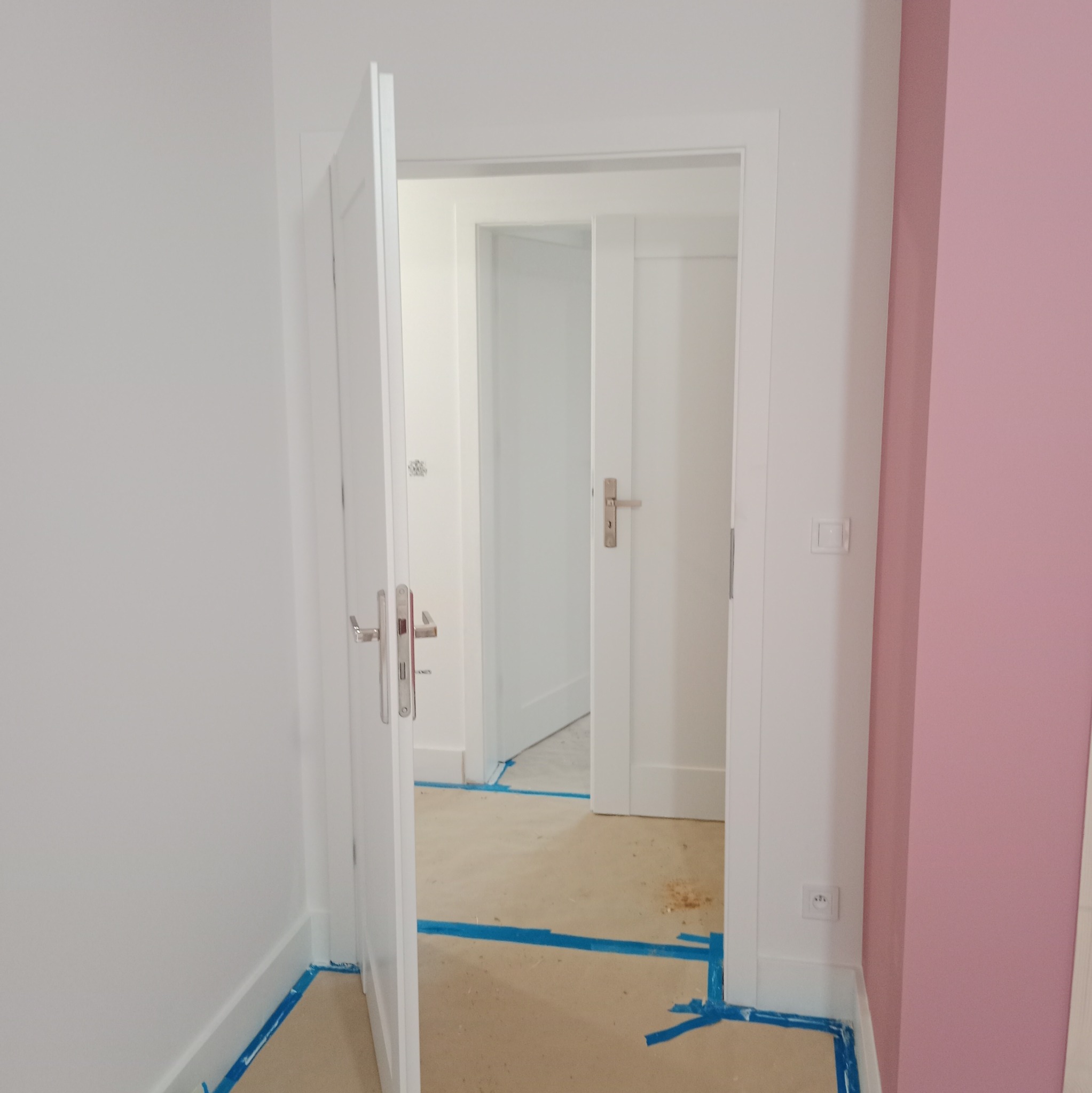 Wnętrze mieszkania w trakcie odświeżania, z otwartymi białymi drzwiami, podłoga zabezpieczona niebieską taśmą malarską, ściany pomalowane na biało i różowo.