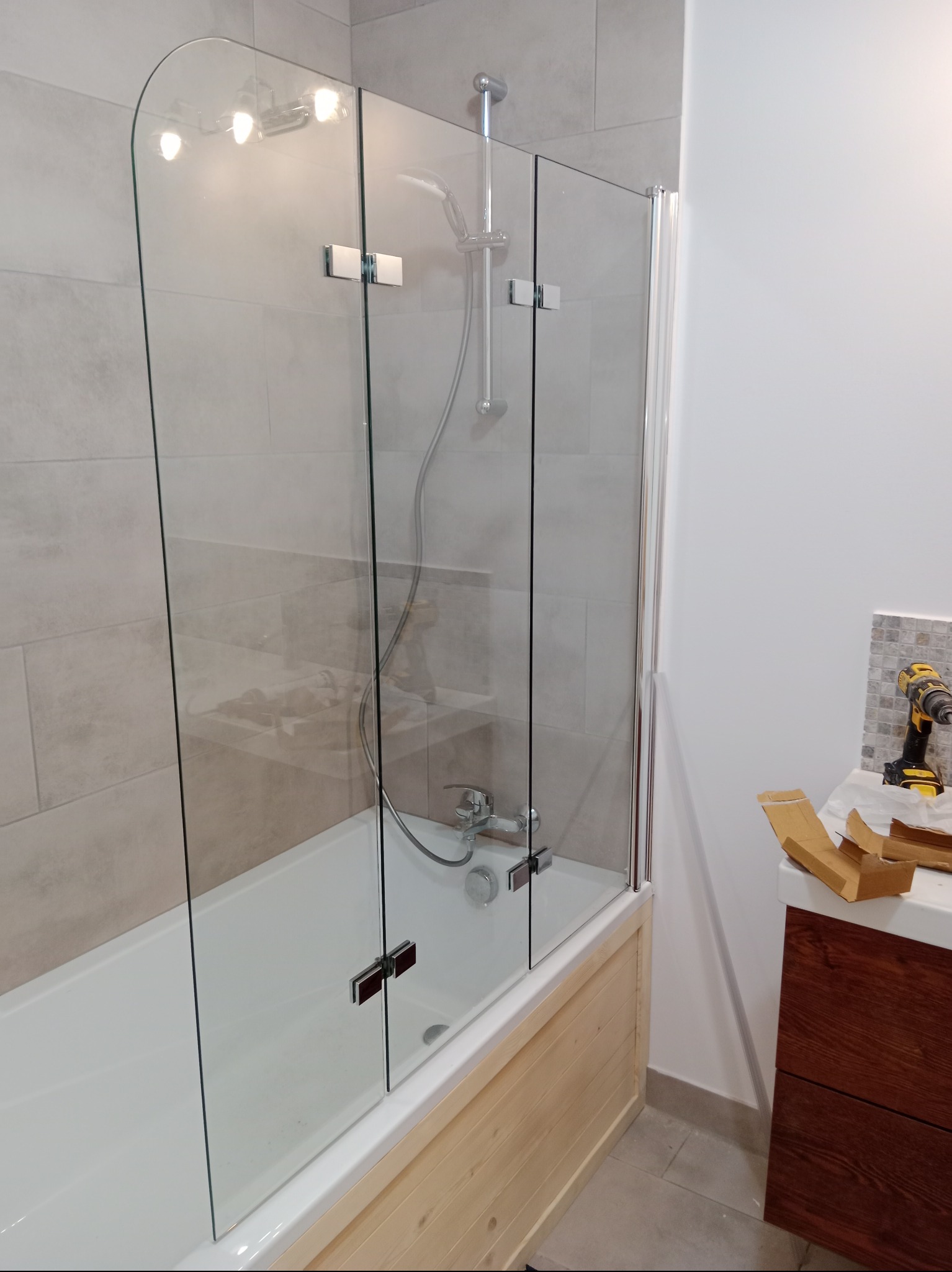 Szklany parawan nawannowy z chromowanymi zawiasami zamontowany nad wanną obudowaną jasnym drewnem, w tle beżowe płytki i fragment szafki z umywalką oraz wiertarka akumulatorowa.