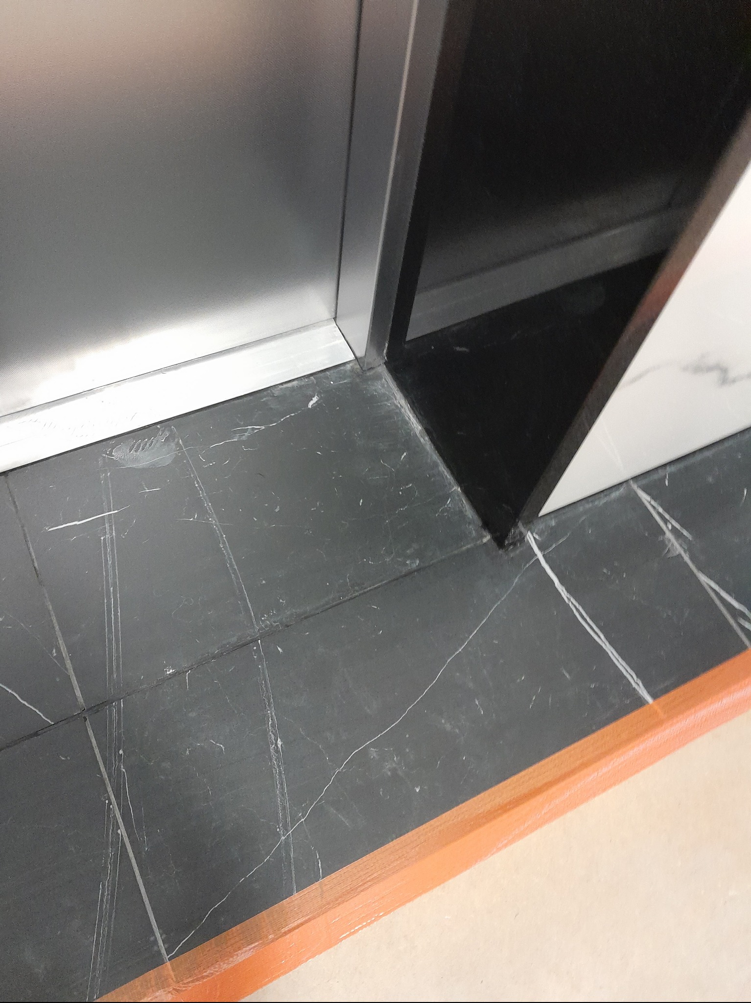 Fragment łazienki z czarną marmurową podłogą, białą marmurową ścianą i srebrną kabiną prysznicową, zabezpieczoną pomarańczową taśmą malarską.