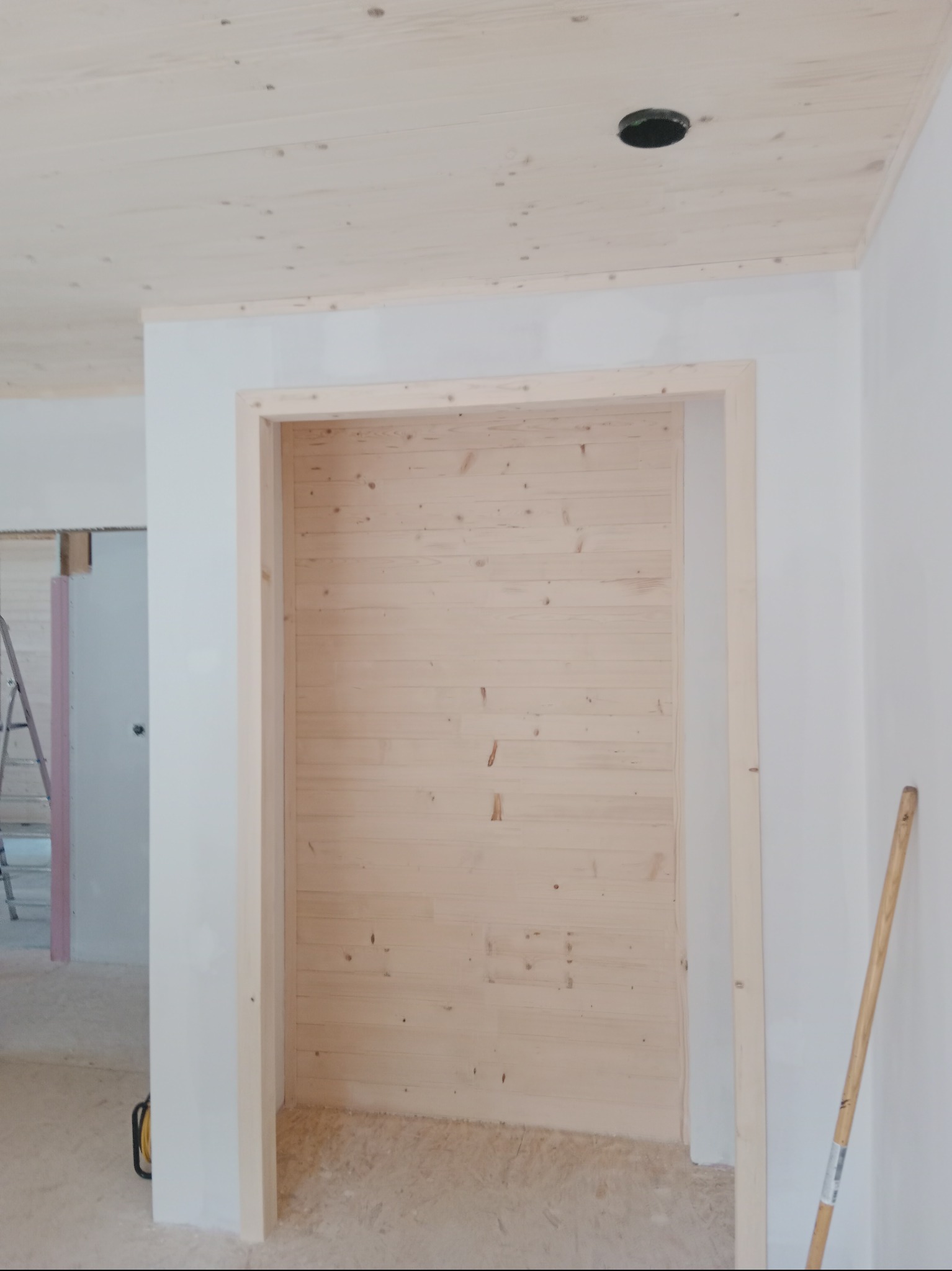 Wnętrze w trakcie budowy domu szkieletowego, widoczna drewniana boazeria na ścianie, surowy sufit z otworem na oświetlenie, białe ściany i fragment drabiny w tle.