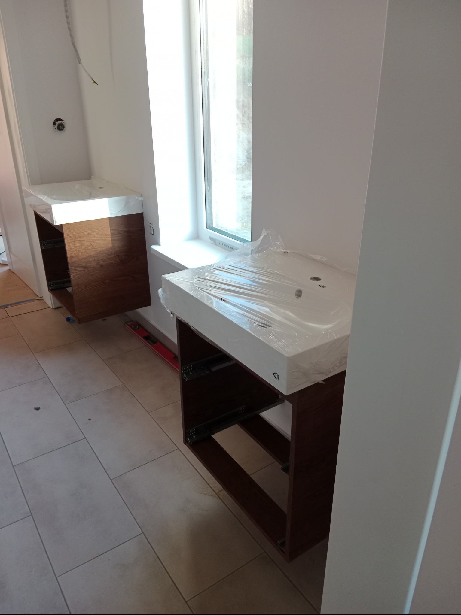 Dwie szafki łazienkowe z umywalkami, jeszcze w folii ochronnej, zamontowane w surowym wnętrzu łazienki z jasnymi płytkami podłogowymi i widocznym poziomnicą.