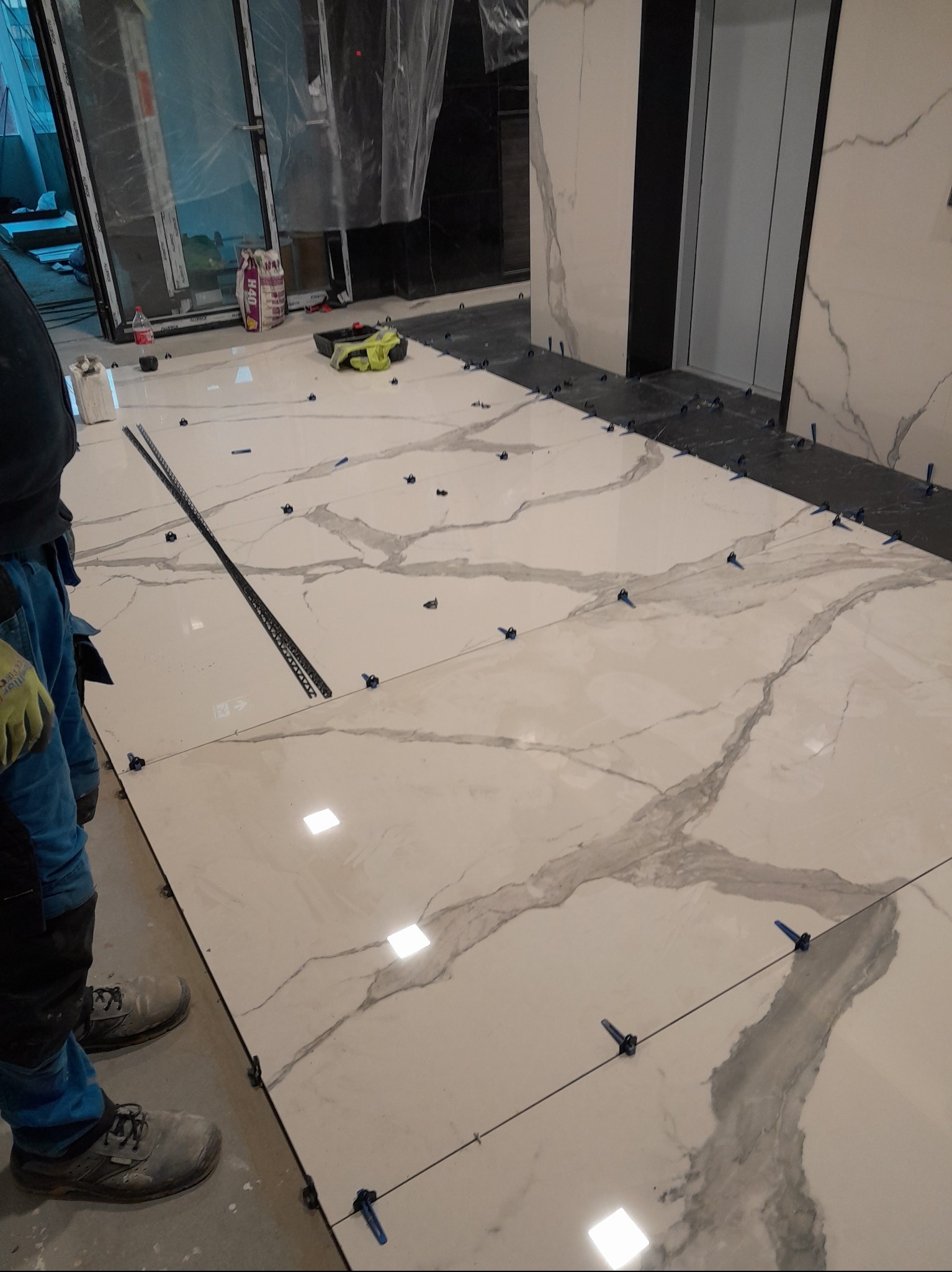 Układanie wielkoformatowych płytek podłogowych z wzorem marmuru, z użyciem systemu poziomowania płytek, widoczny pracownik w roboczym ubraniu i narzędzia.