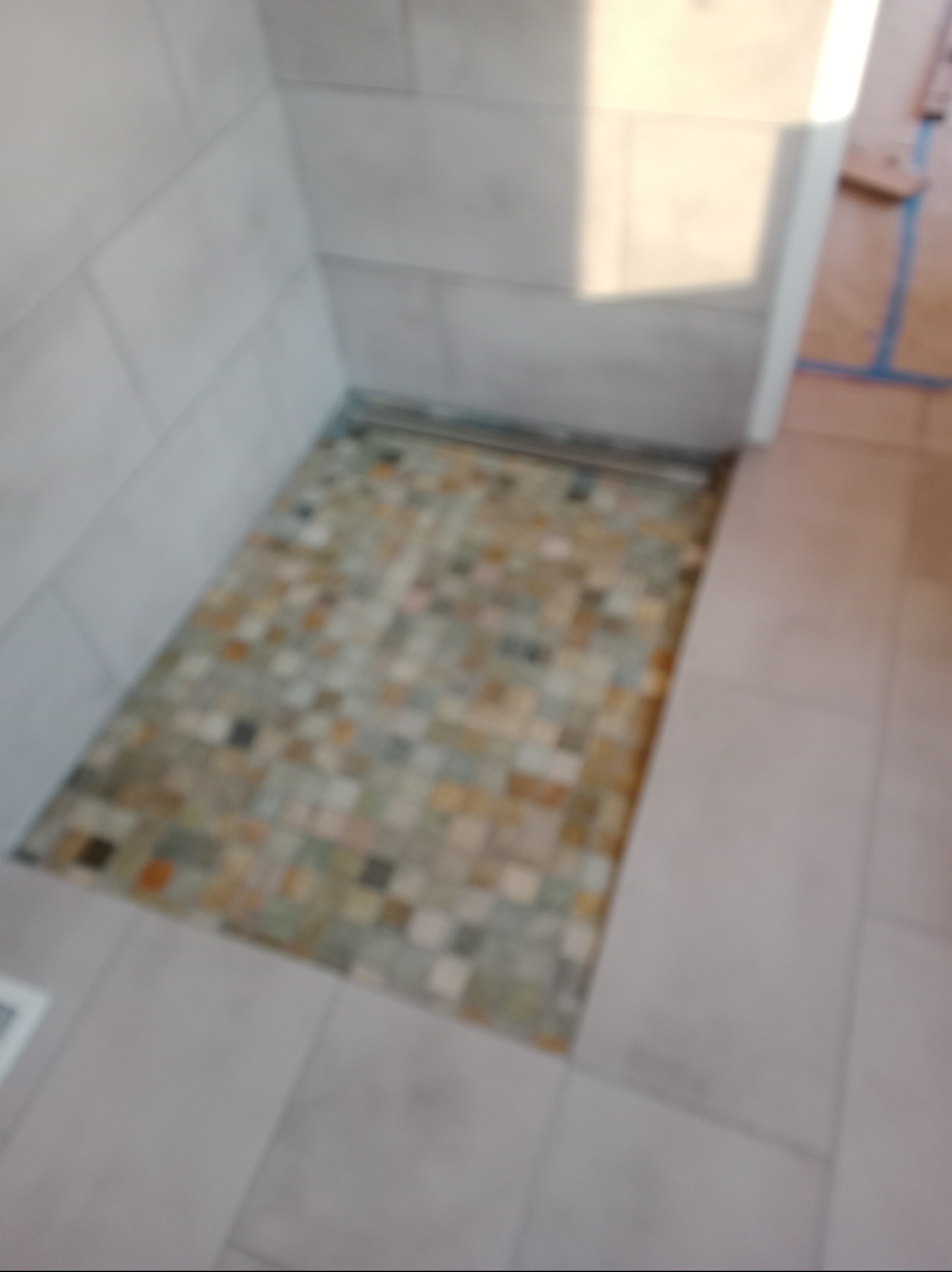 Ułożona mozaika z drobnych płytek w brodziku kabiny prysznicowej, otoczona jasnymi, kwadratowymi płytkami ściennymi i podłogowymi.